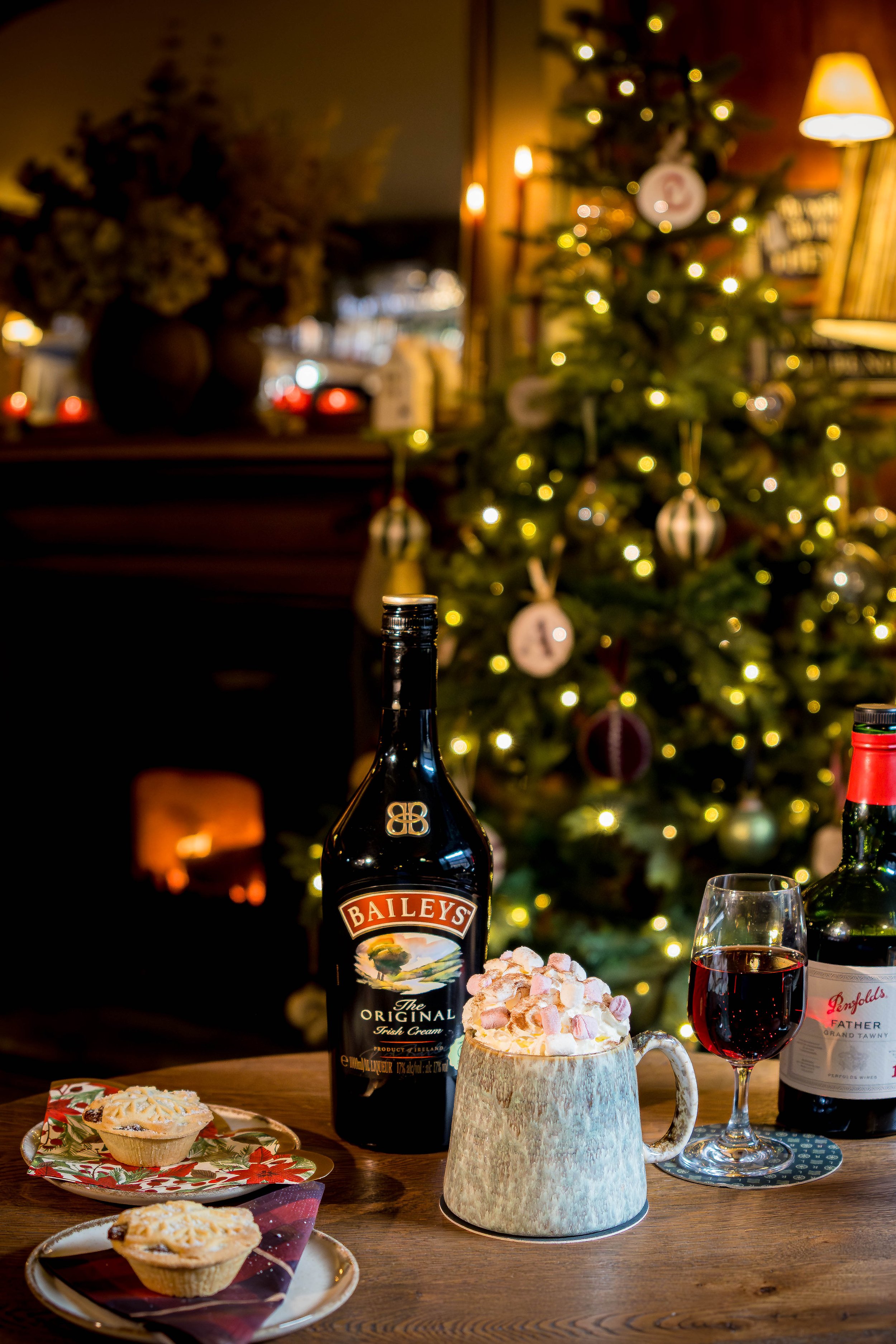 Christmas scene with a decorated tree, a bottle of Baileys Irish Cream, a glass of red wine, a mug of hot chocolate topped with marshmallows, and holiday treats on a wooden table.