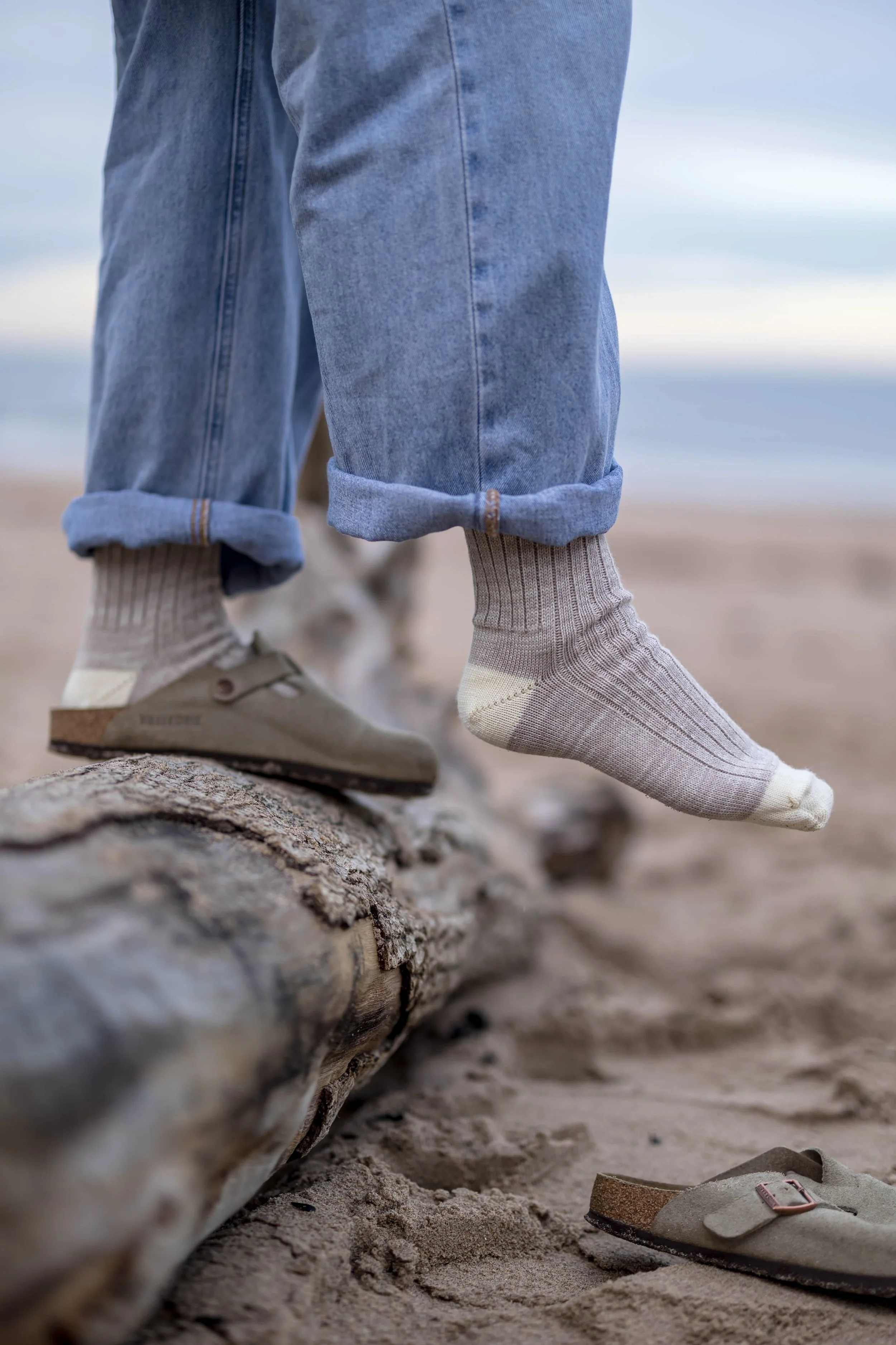 Person walking on a beach, with rolled-up jeans, one foot in a sneaker, and the other foot in a sock, with a flip-flop sandal on the sand nearby.
