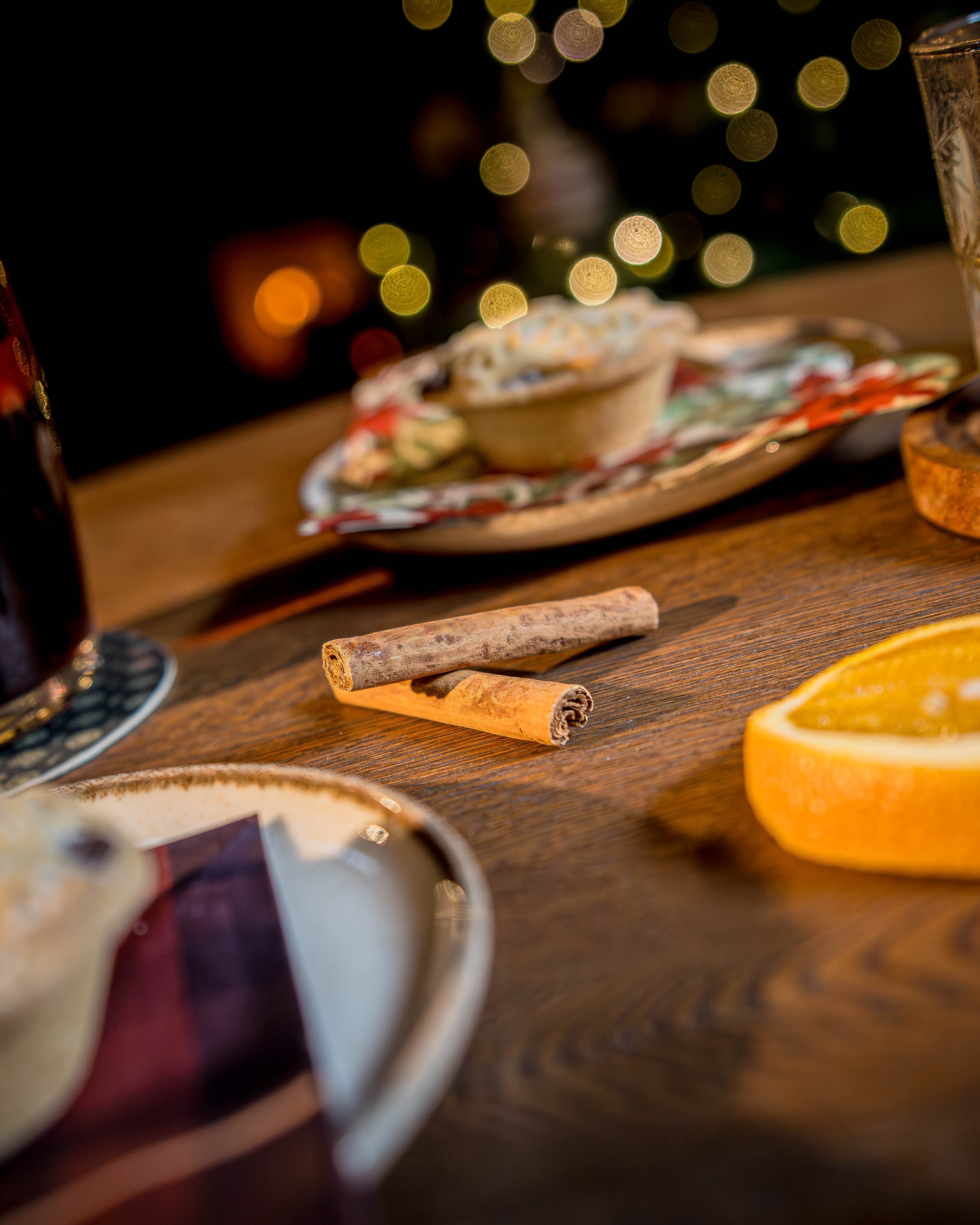 Close-up of cinnamon sticks on a wooden table with a slice of orange, out-of-focus Christmas lights in the background, and holiday-themed dishes nearby.