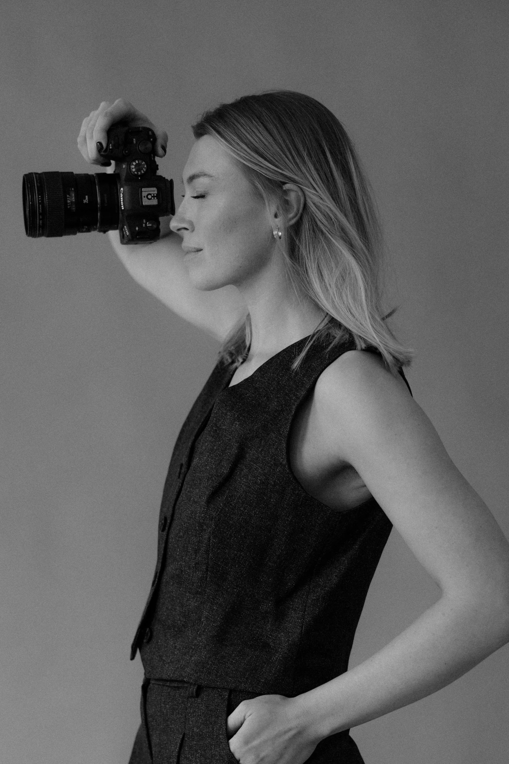 Black and white photo of a woman with shoulder-length hair, wearing a sleeveless vest, holding a camera up to her eye with her right hand, and standing with her left hand in her pocket, with her eyes closed, against a plain background.