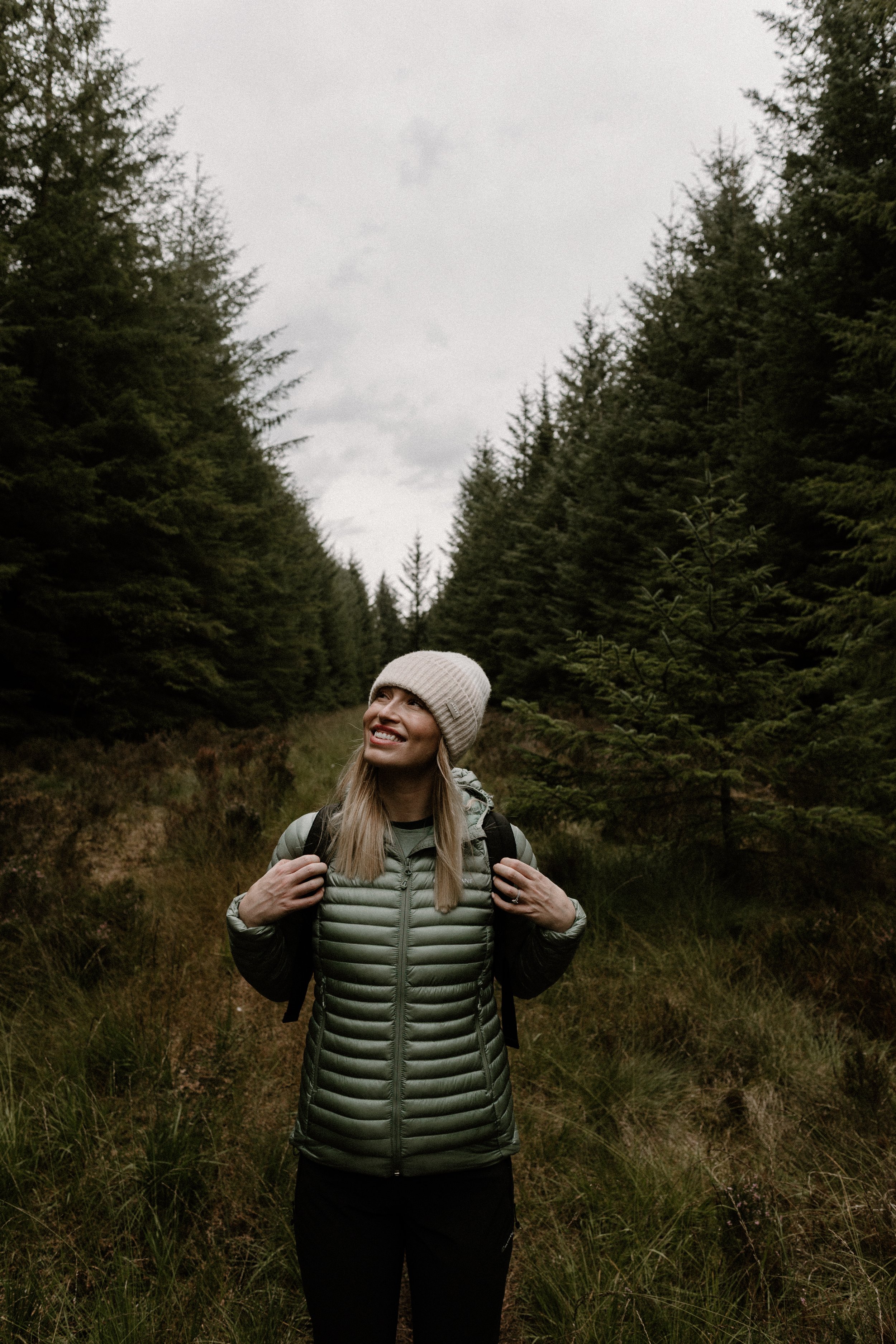 A woman in outdoor gear and a beanie hiking through a forested trail, smiling and looking up.