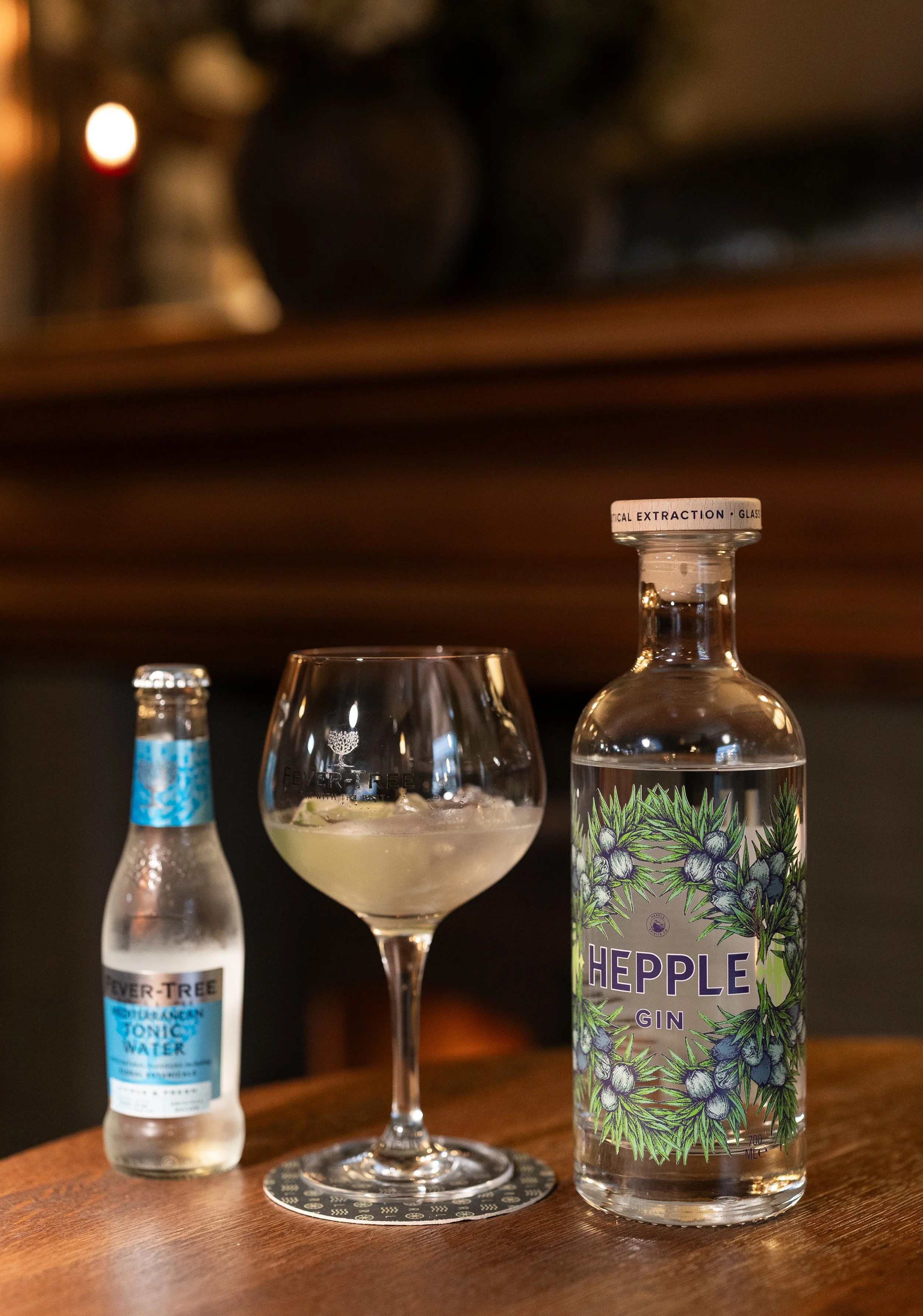 A bottle of Hepple gin, a tonic water bottle, and a drink in a glass on a wooden surface.