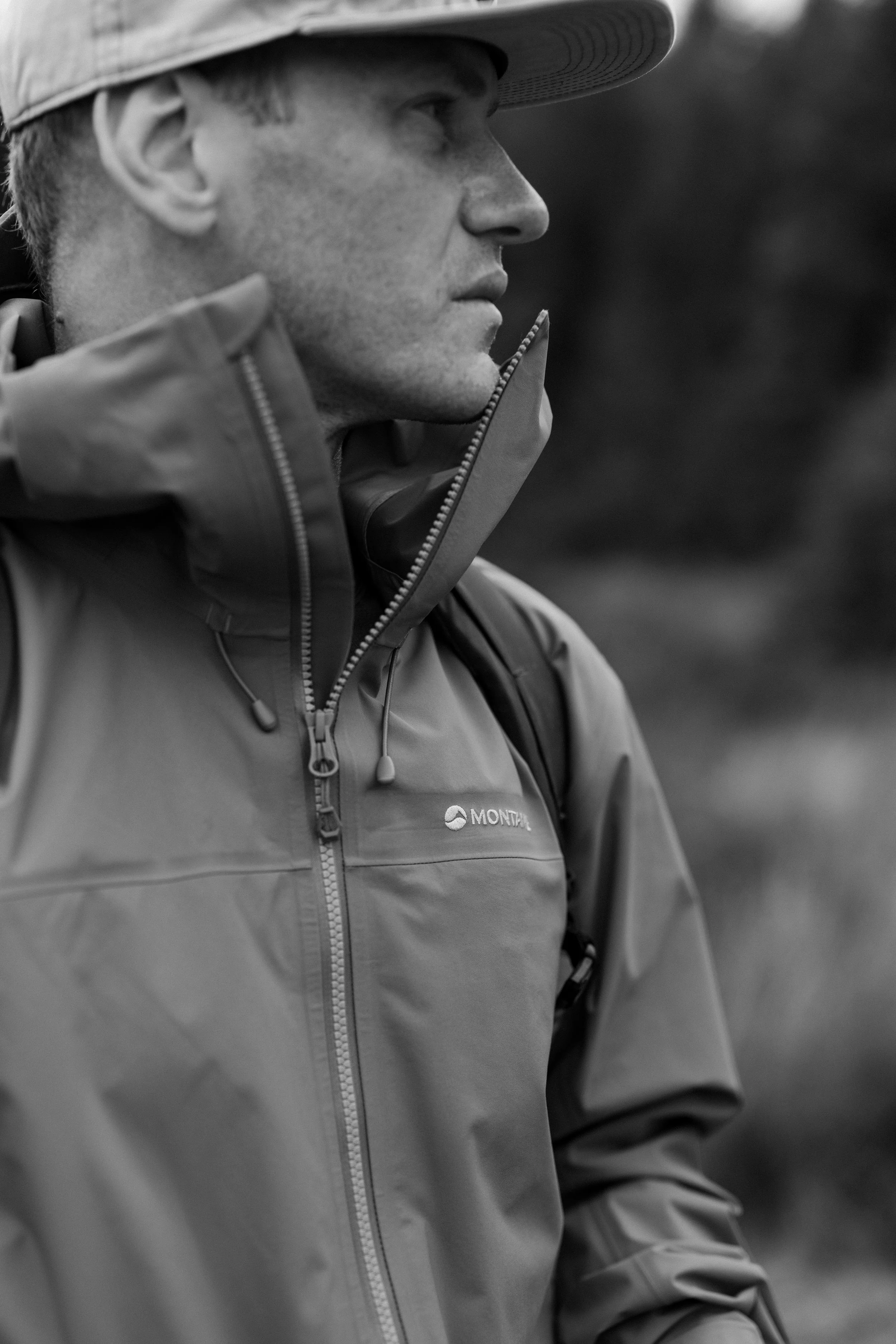 A man wearing a wide-brim hat and outdoor jacket looking to the right outdoors.