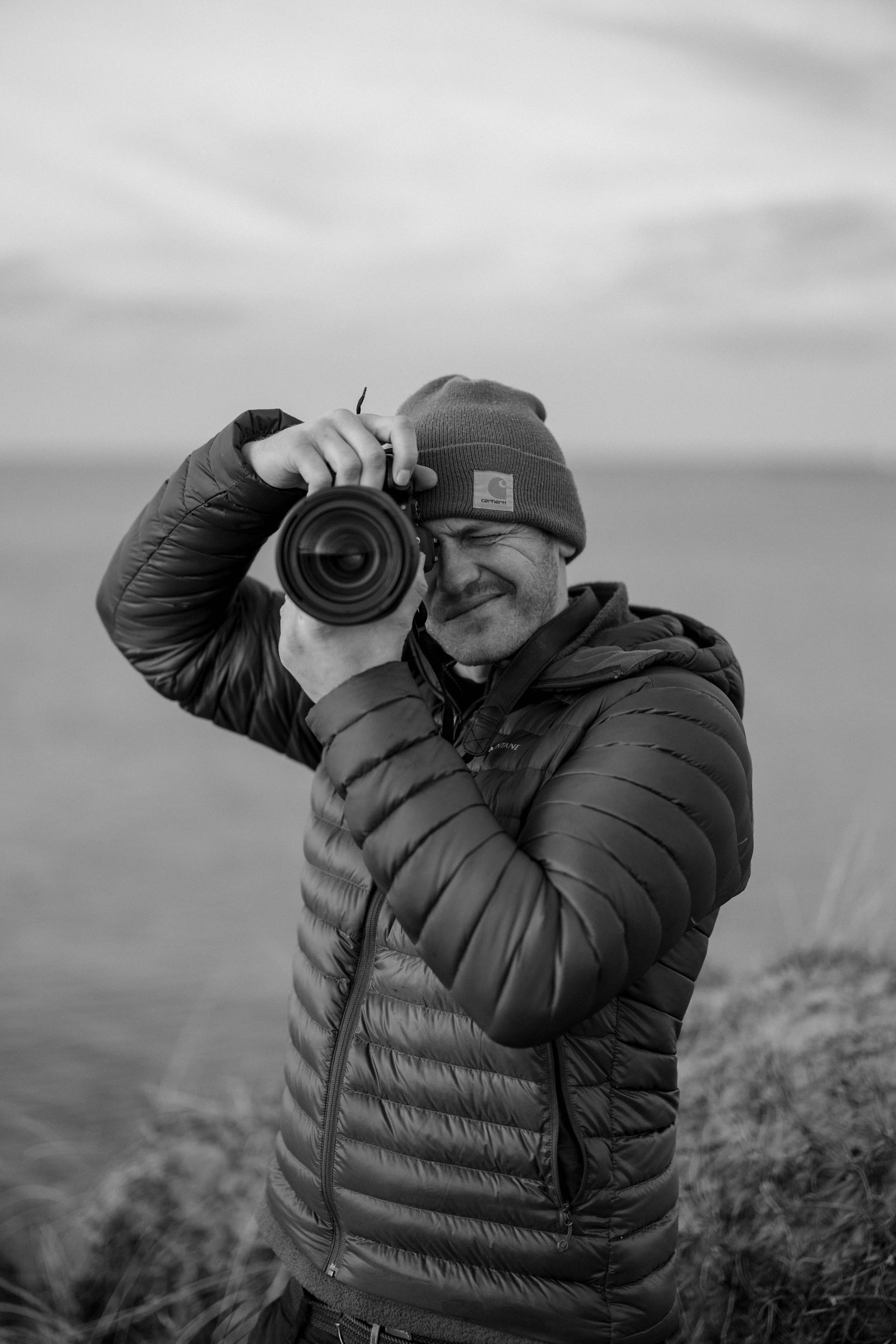 A man in a puffy jacket and beanie hat taking a photograph outdoors with a camera, squinting and smiling, standing on a grassy hill with a cloudy sky in the background.