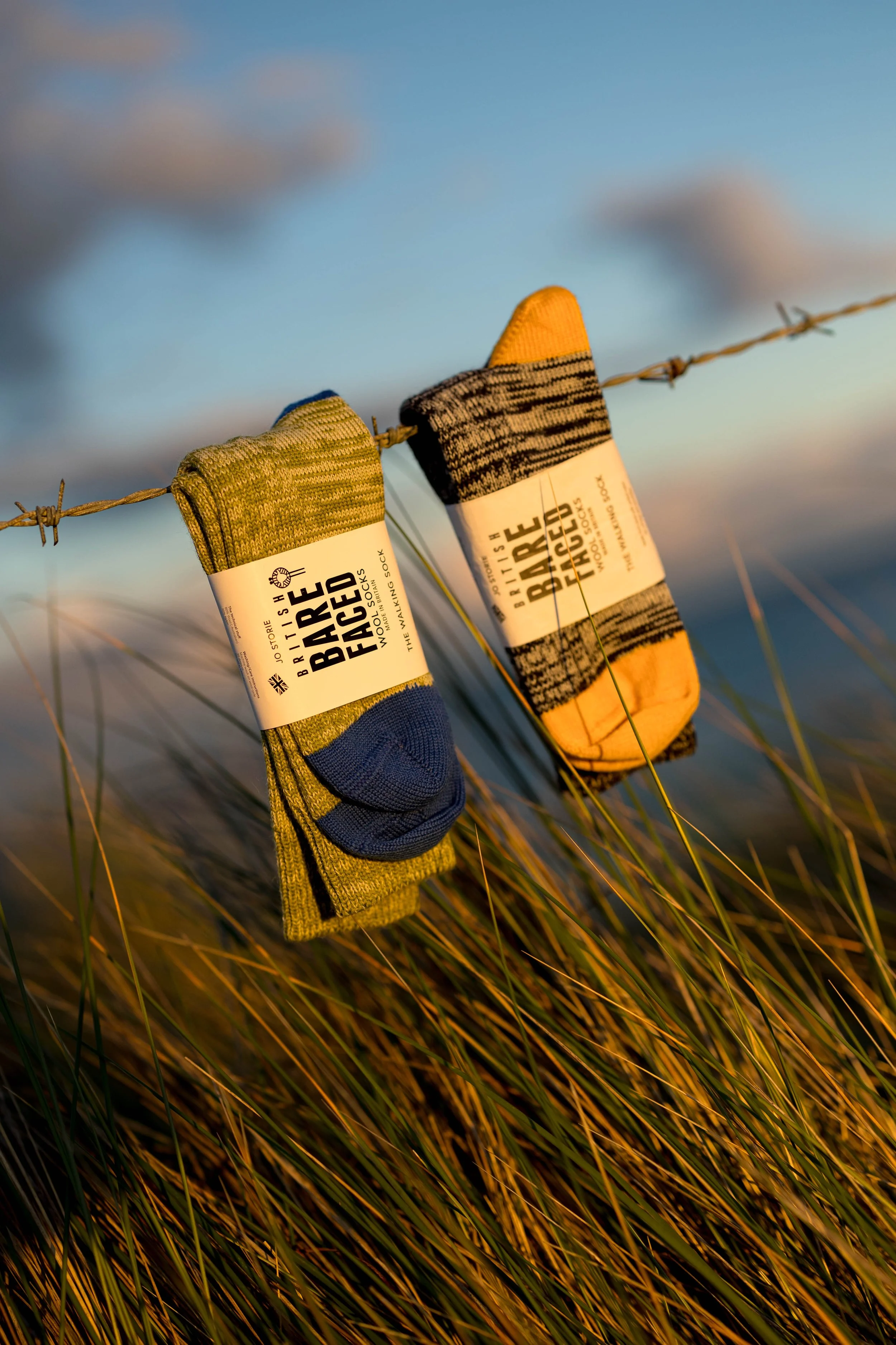Two pairs of colorful socks hanging on a barbed wire fence in a grassy field with a cloudy sky background.
