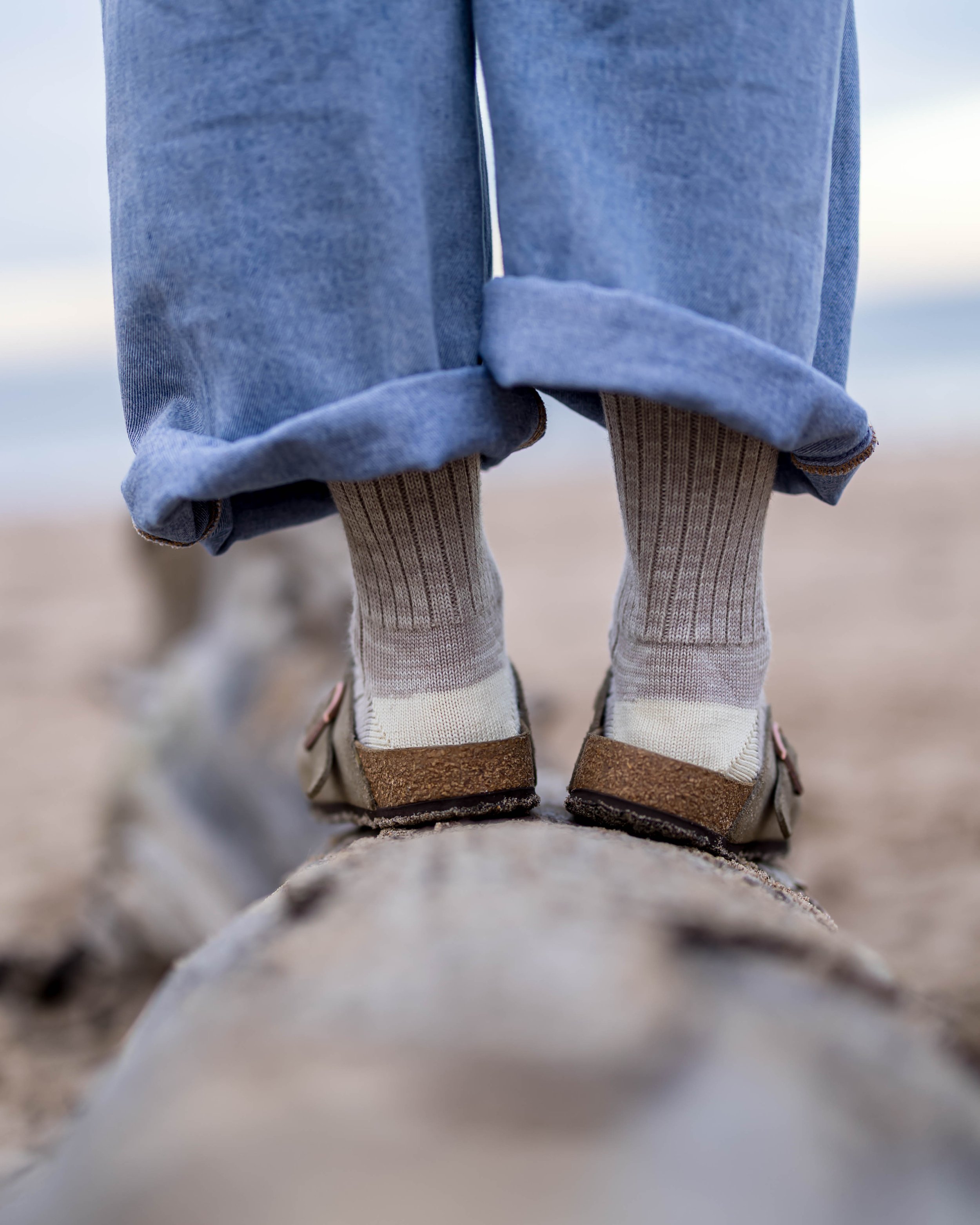 Close-up of a person's feet standing on a log on the beach, with rolled-up jeans and socks, and sandals.