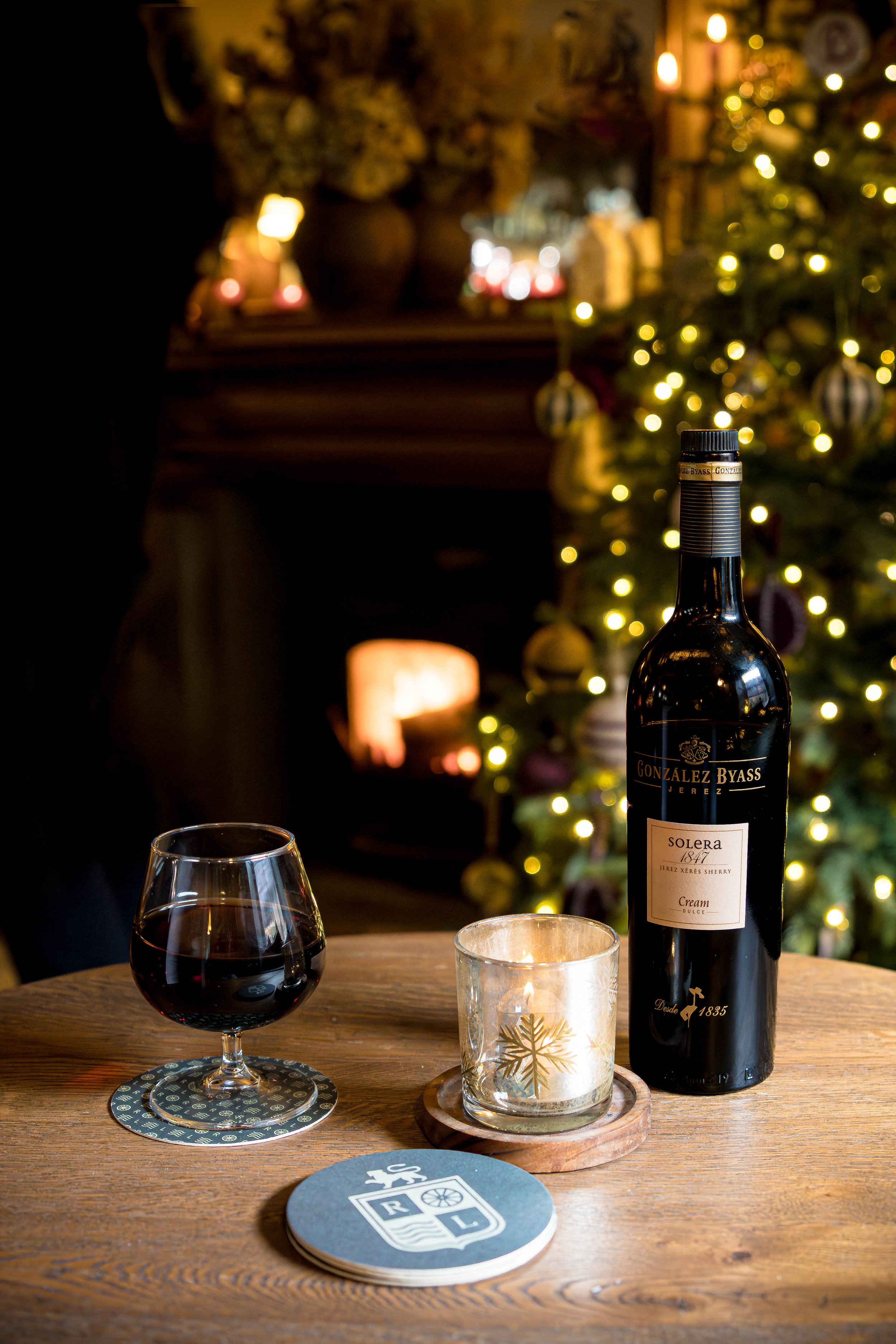 A table with a glass of red wine, a lit candle in a glass holder, a bottle of Gonzalez Byass Solera wine, and coasters with decorative designs, set in a cozy room decorated for Christmas with a Christmas tree and a lit fireplace in the background.