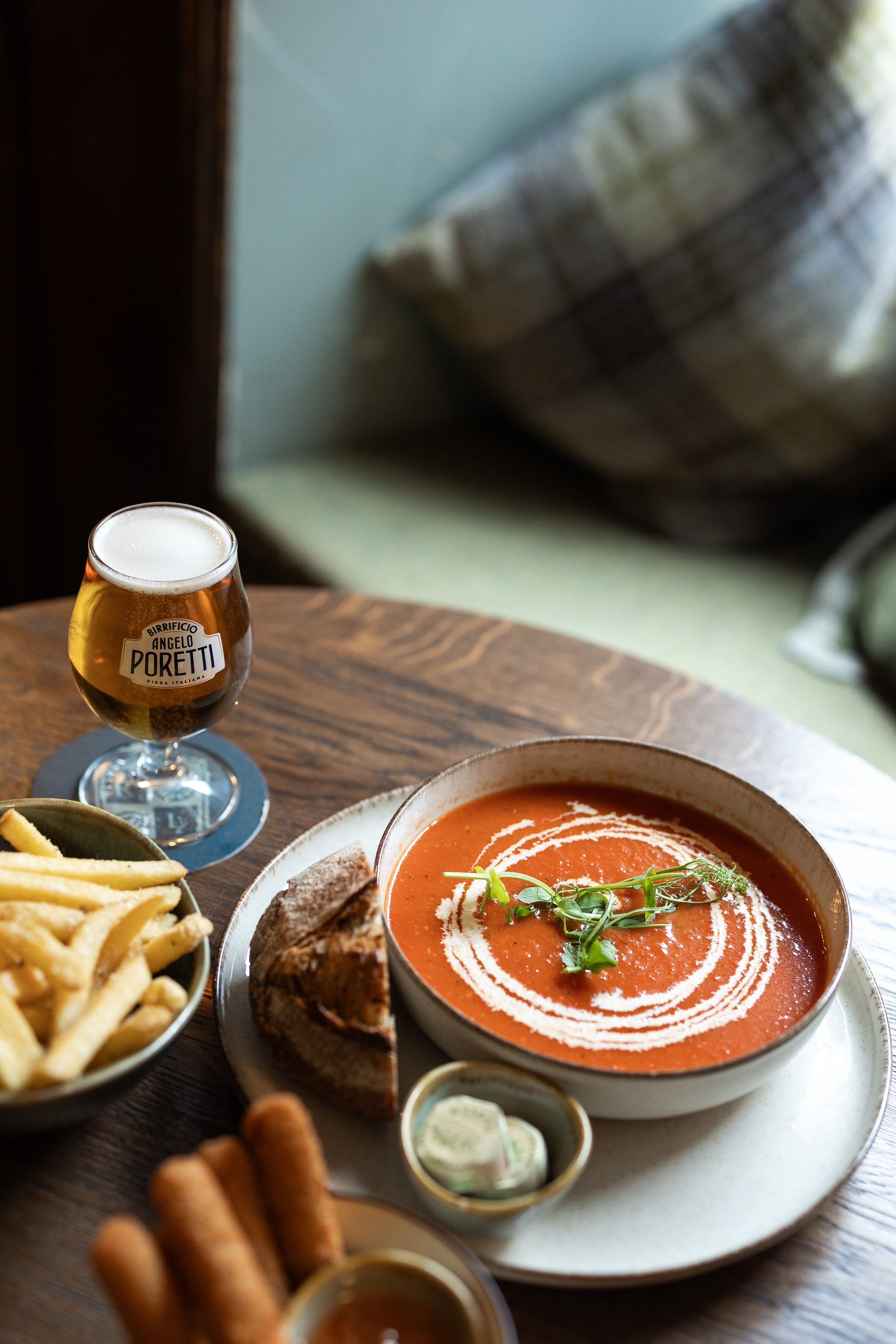 A bowl of tomato soup garnished with greens, a half pizza, a bowl of French fries, and a glass of beer on a wooden table.