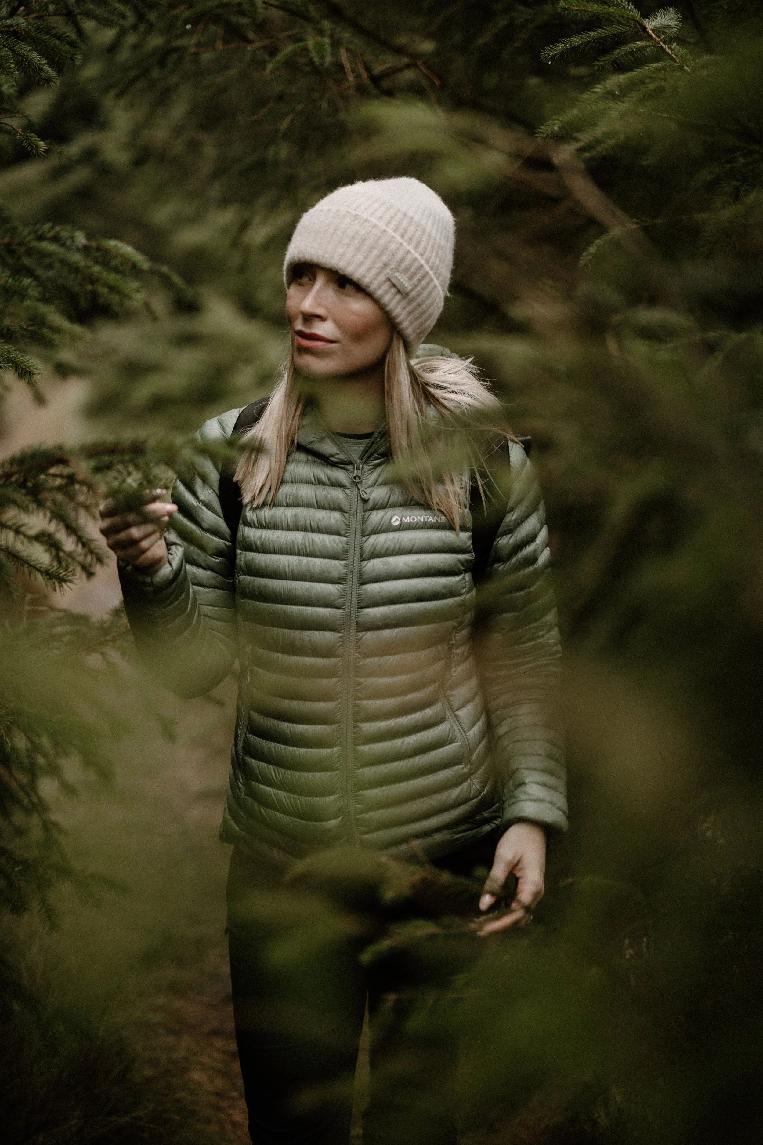 A woman in outdoor gear, including a beige knit hat and a green puffer jacket, walking through a forested area with evergreen trees.