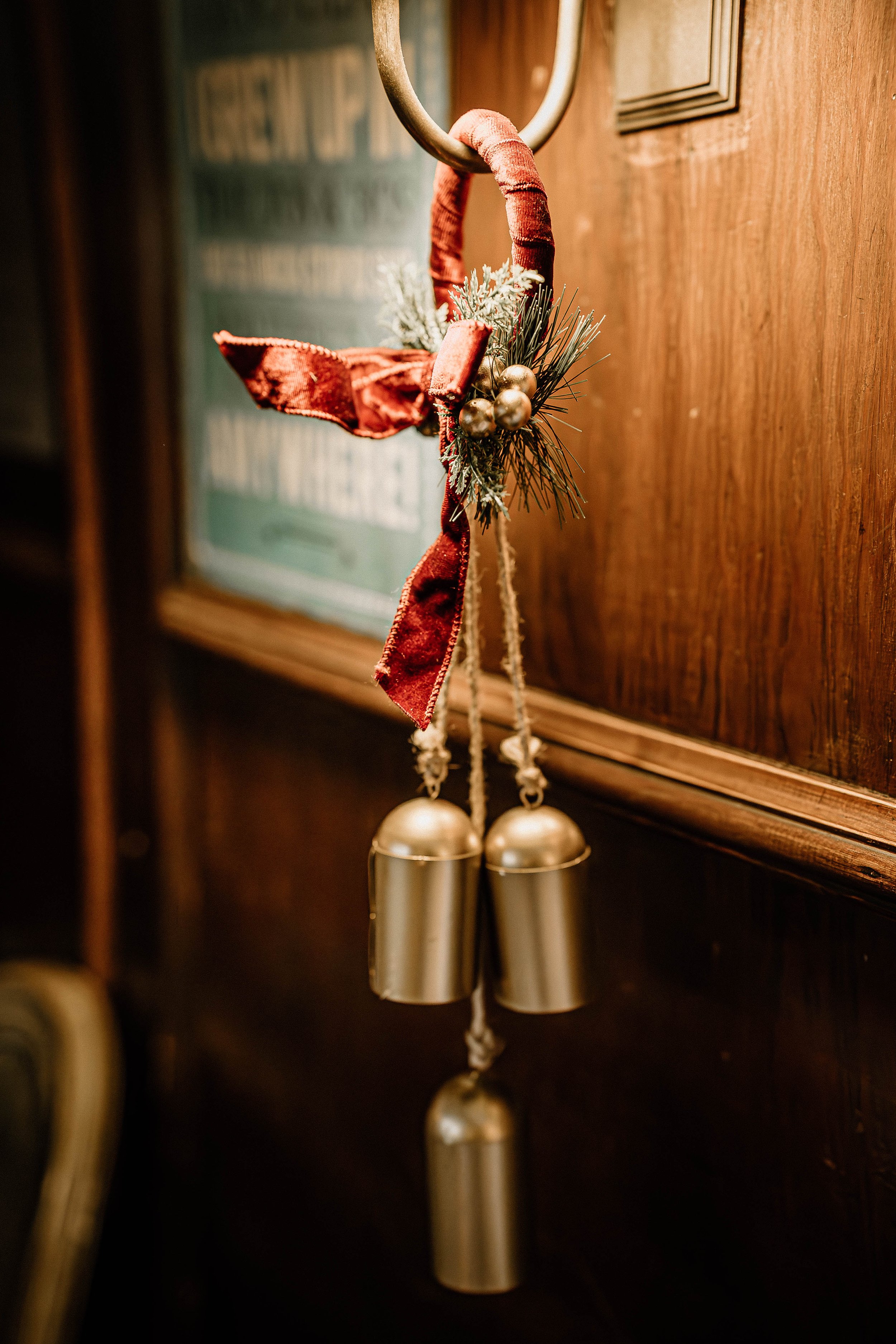 A festive Christmas bell with a red velvet ribbon, pine branches, and gold ornaments hanging on a wooden wall.