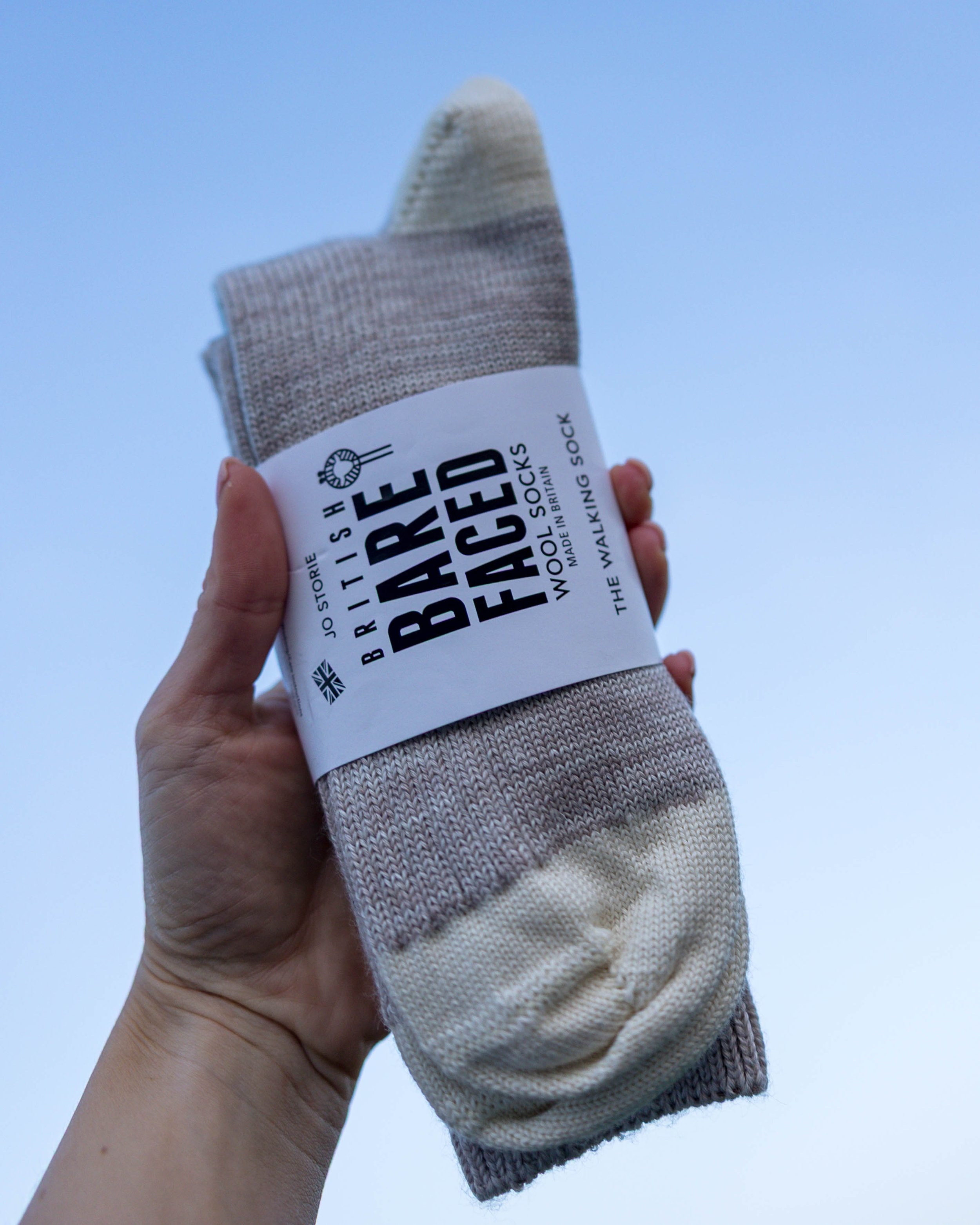 A hand holding a pair of beige and white striped socks with a label that reads "Barefoot British Wool Socks, Made in Britain, The Walking Sock," against a cloudy sky.
