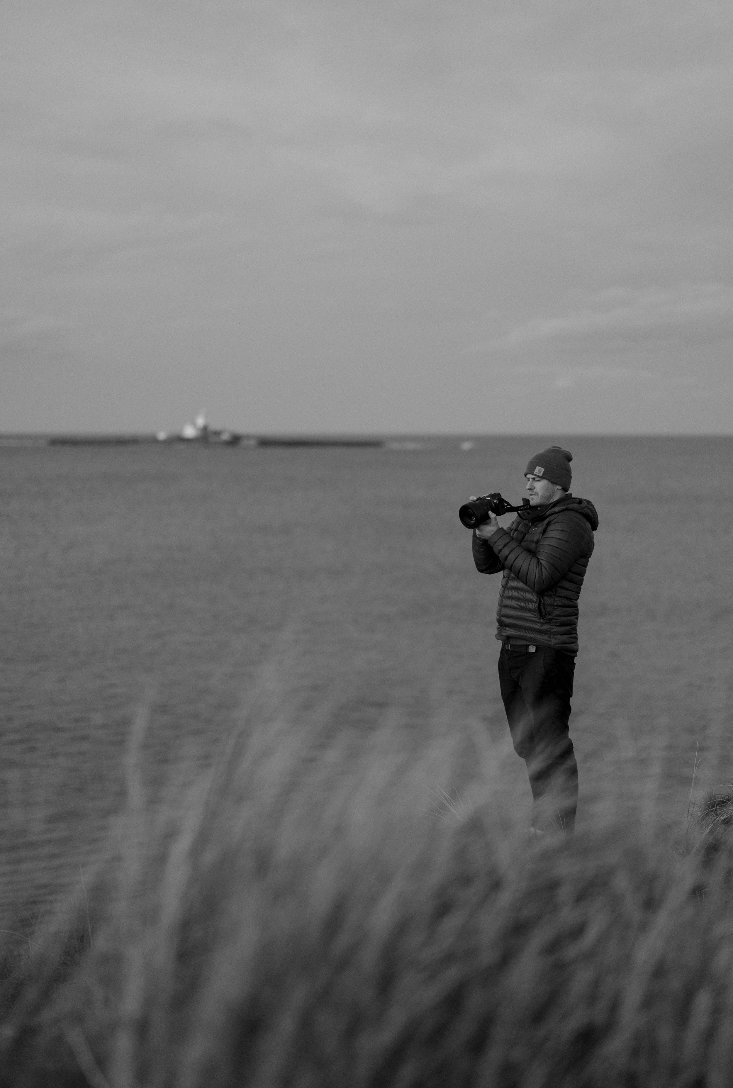 A man standing near the water's edge holding a camera, wearing a beanie and a puffy jacket, with grasses in the foreground and a boat in the-distance.