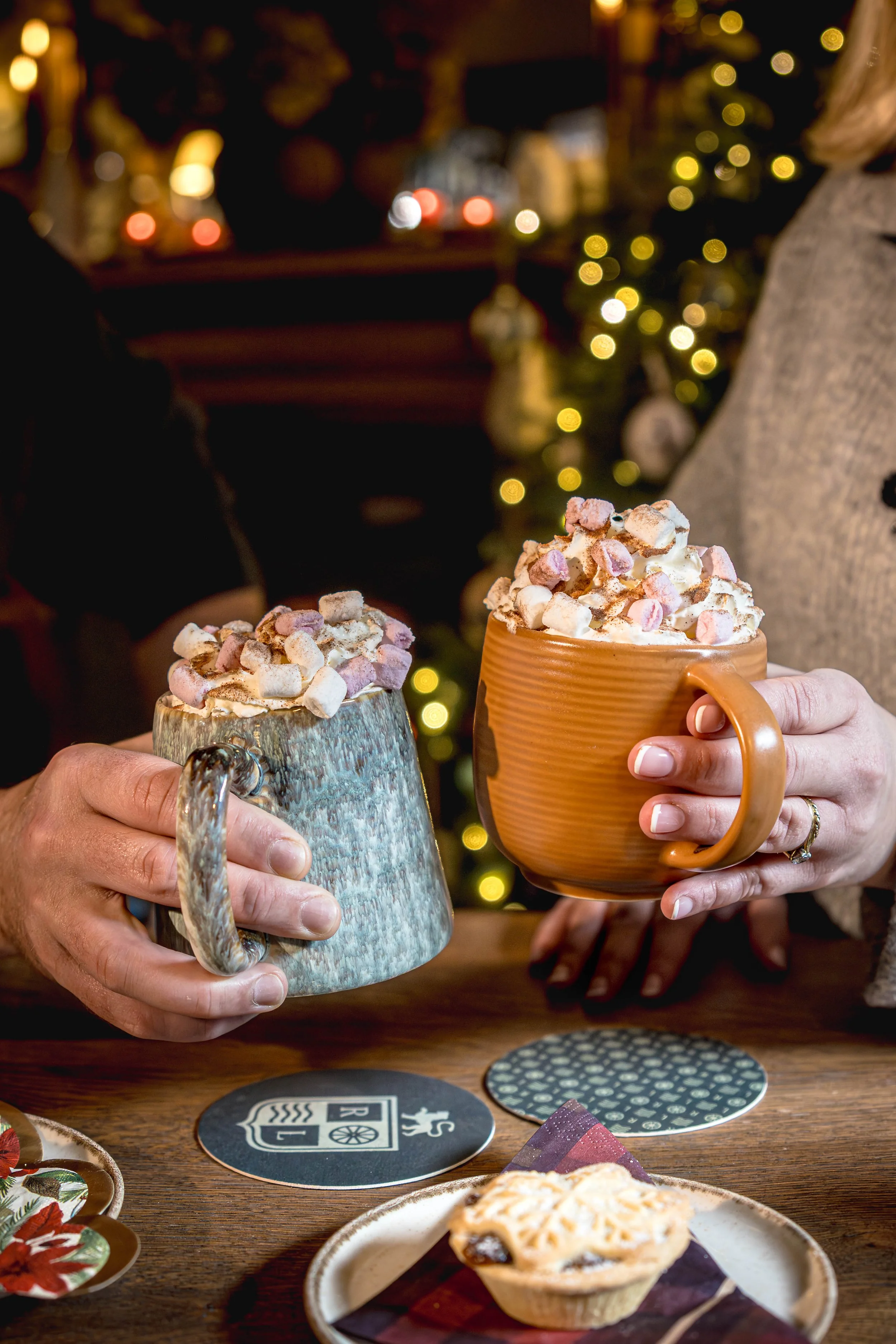 Two people holding mugs filled with hot chocolate topped with marshmallows, with Christmas decorations and a decorated tree in the background.