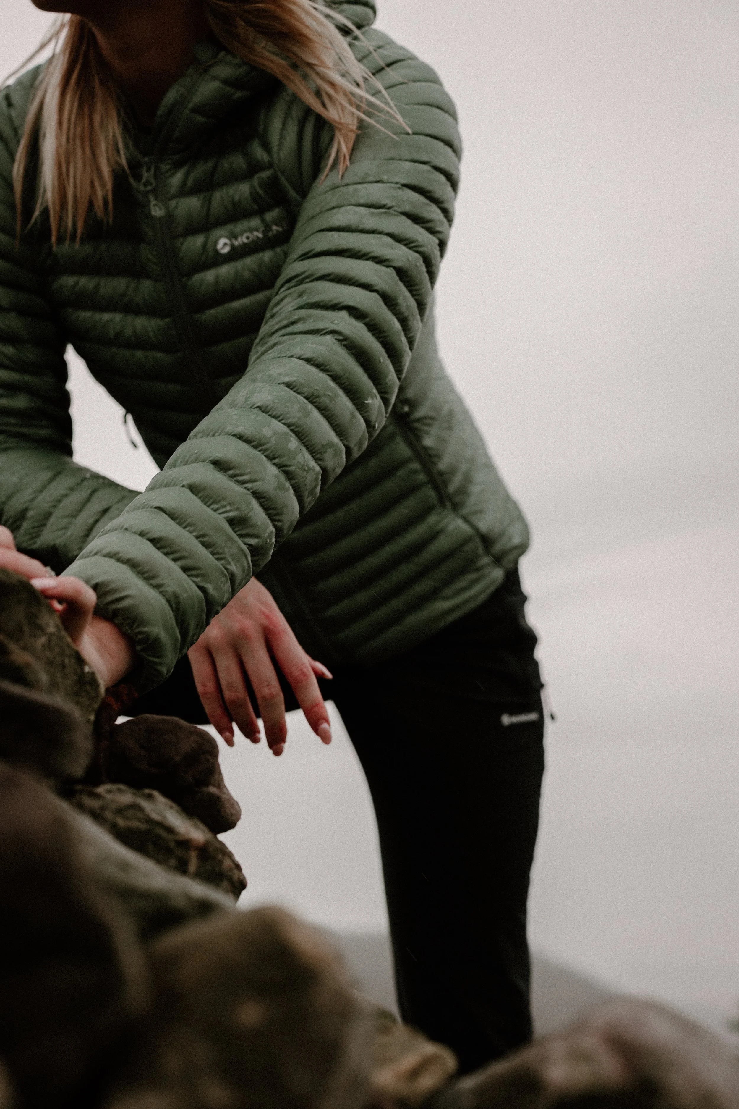Person in a green puffer jacket leaning over and interacting with rocks outdoors on a cloudy day.