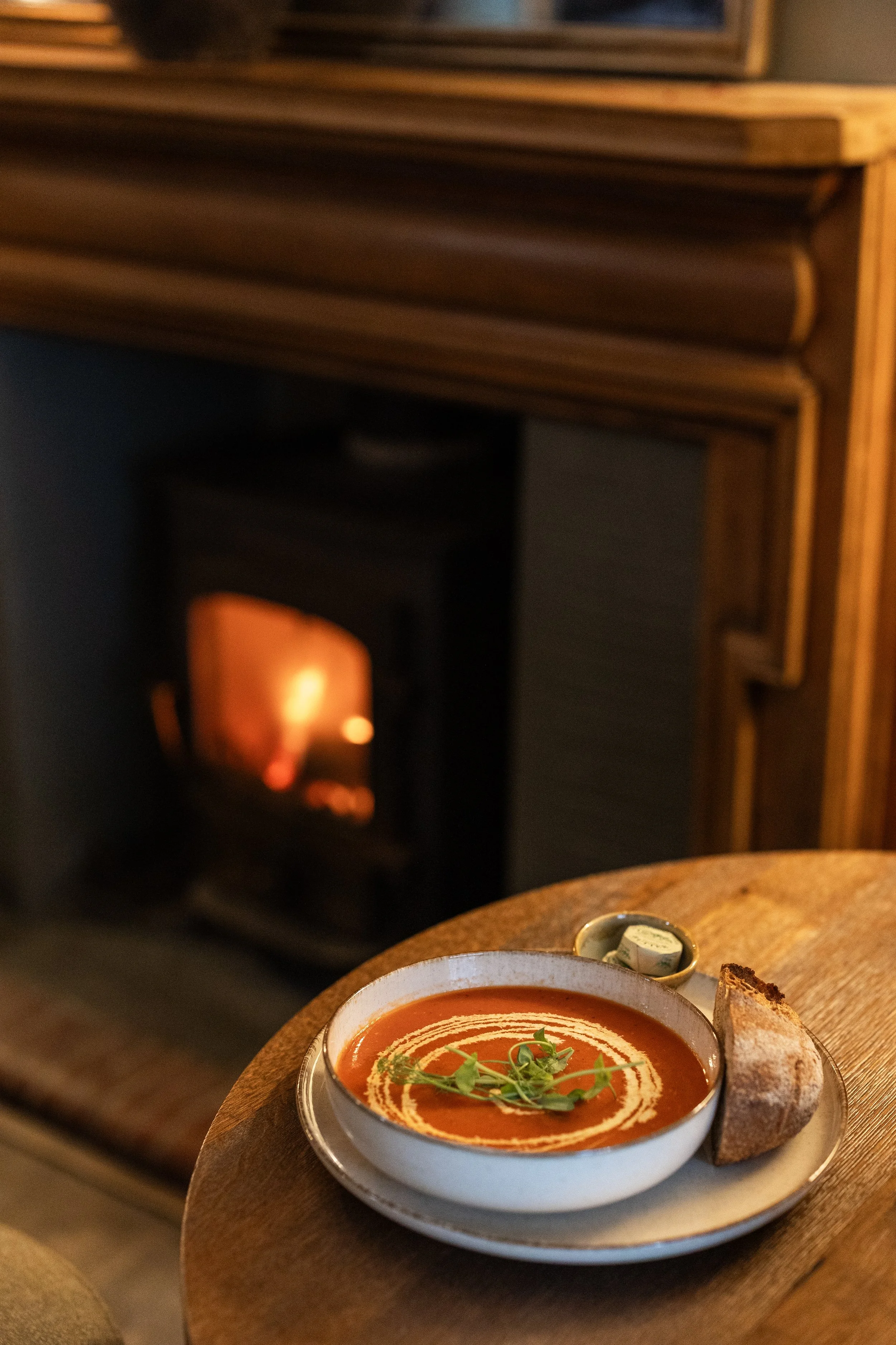 A bowl of tomato soup garnished with cream and microgreens, served with a piece of bread, on a round wooden table in front of a fireplace.