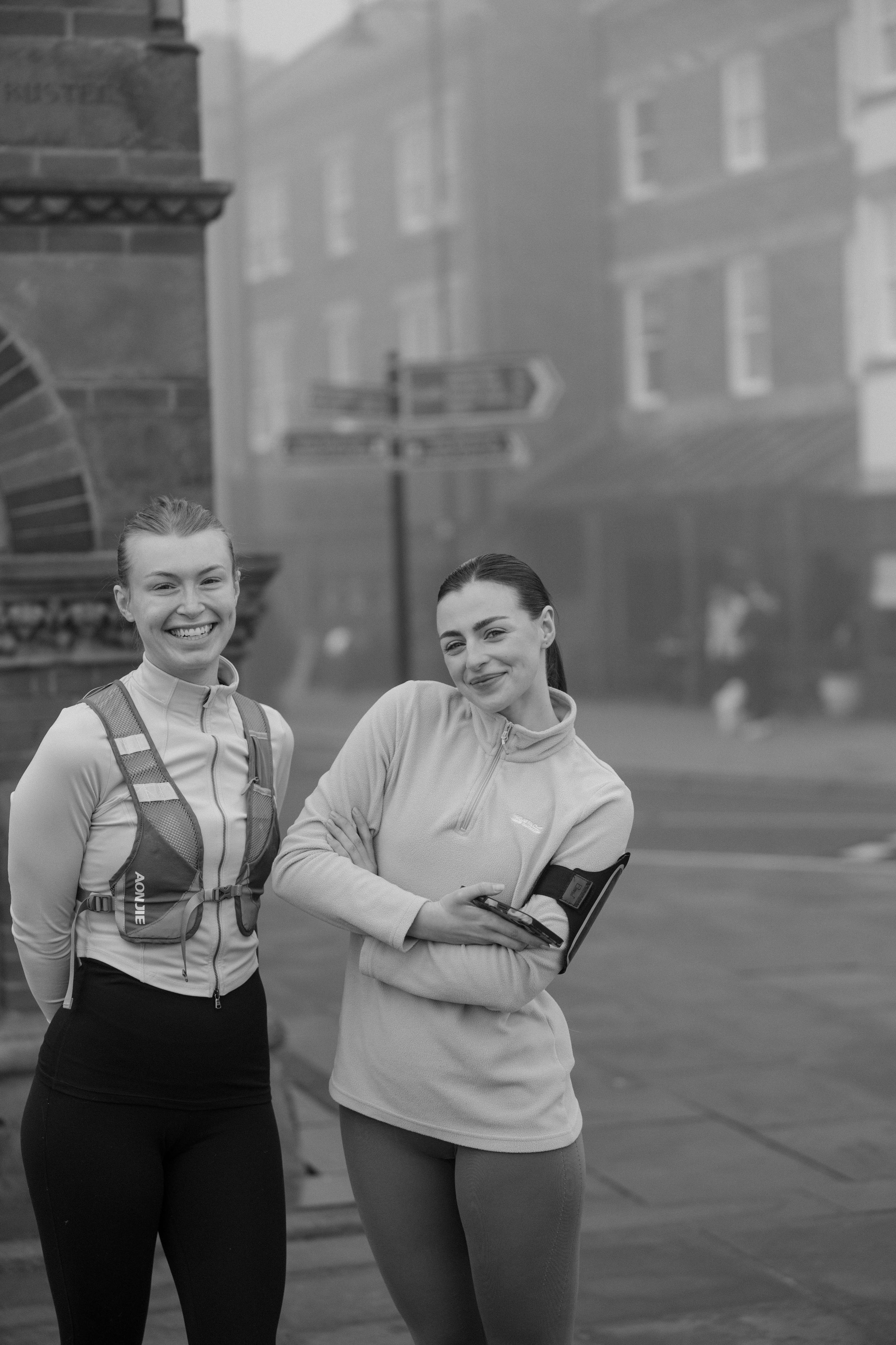 Two women standing outdoors, smiling for the photo. They are dressed in athletic clothing, with one holding a smartphone and wearing a small backpack. The background shows a city street and buildings, with a signpost and some blurred figures.