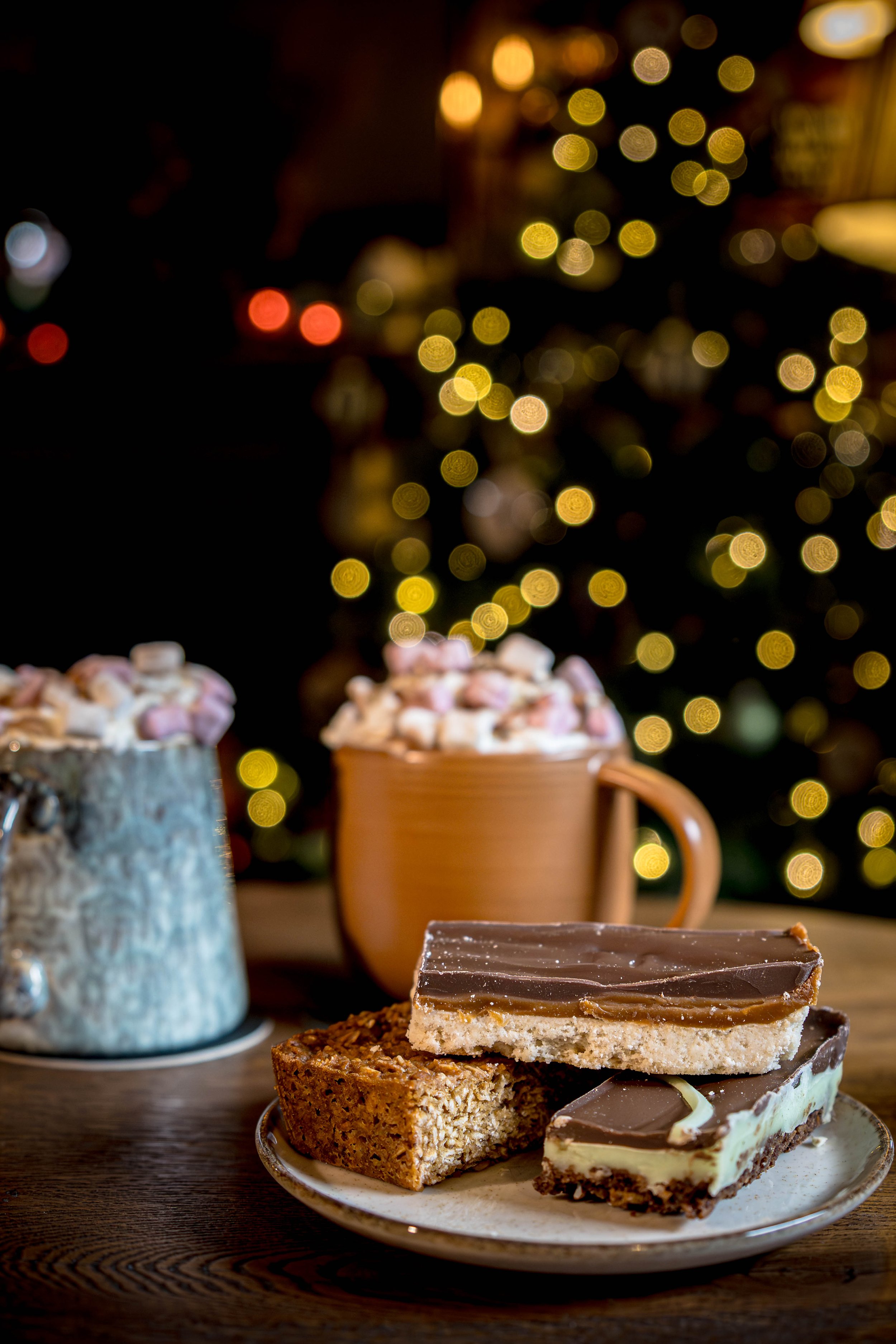 Assorted desserts including chocolate and layered bars on a plate with a background of holiday lights and two mugs of hot chocolate topped with marshmallows.
