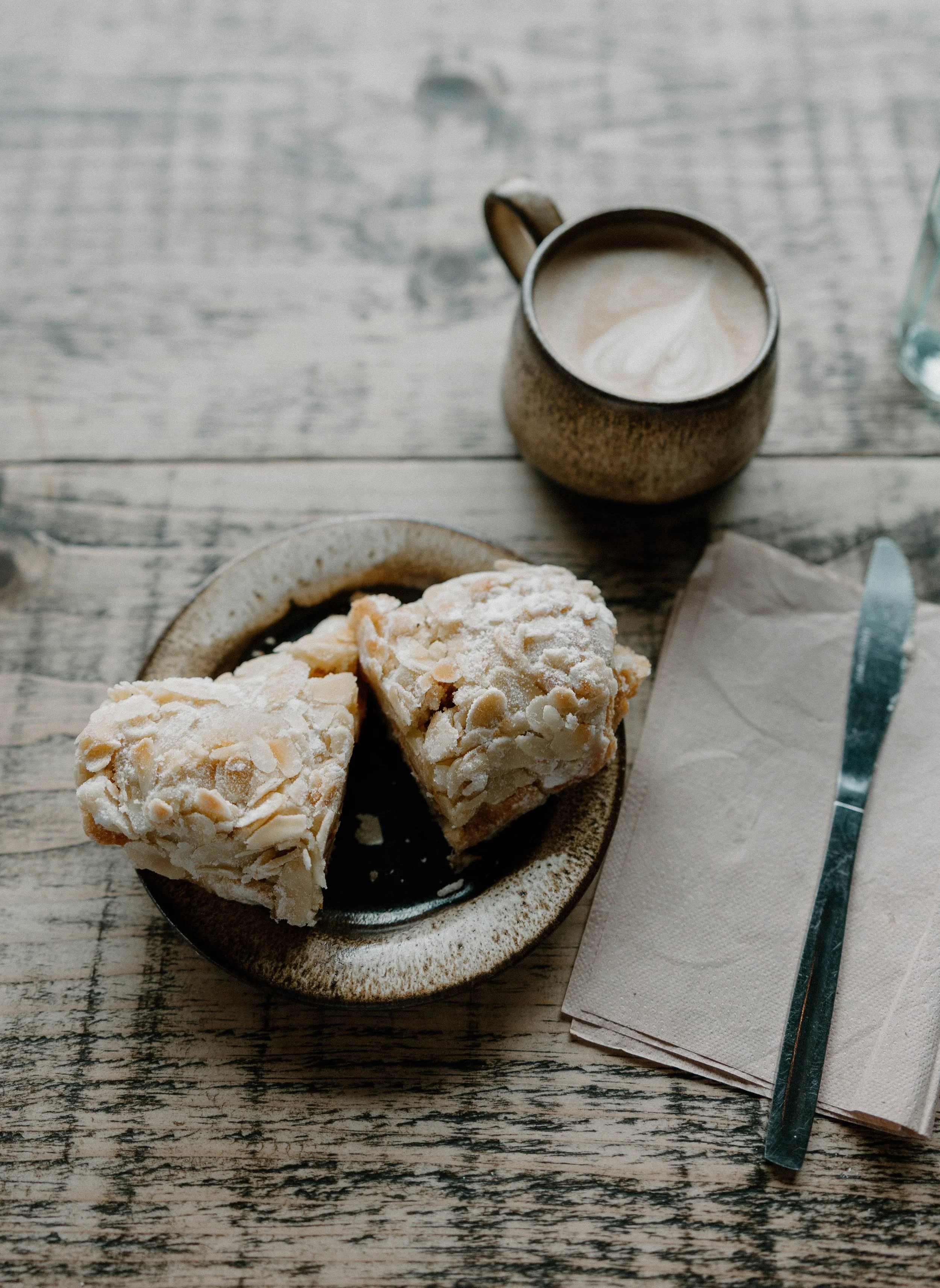 Cookies with almond slices on a plate, a cup of coffee with foam art, a glass of water, a napkin, and a knife on a wooden table.