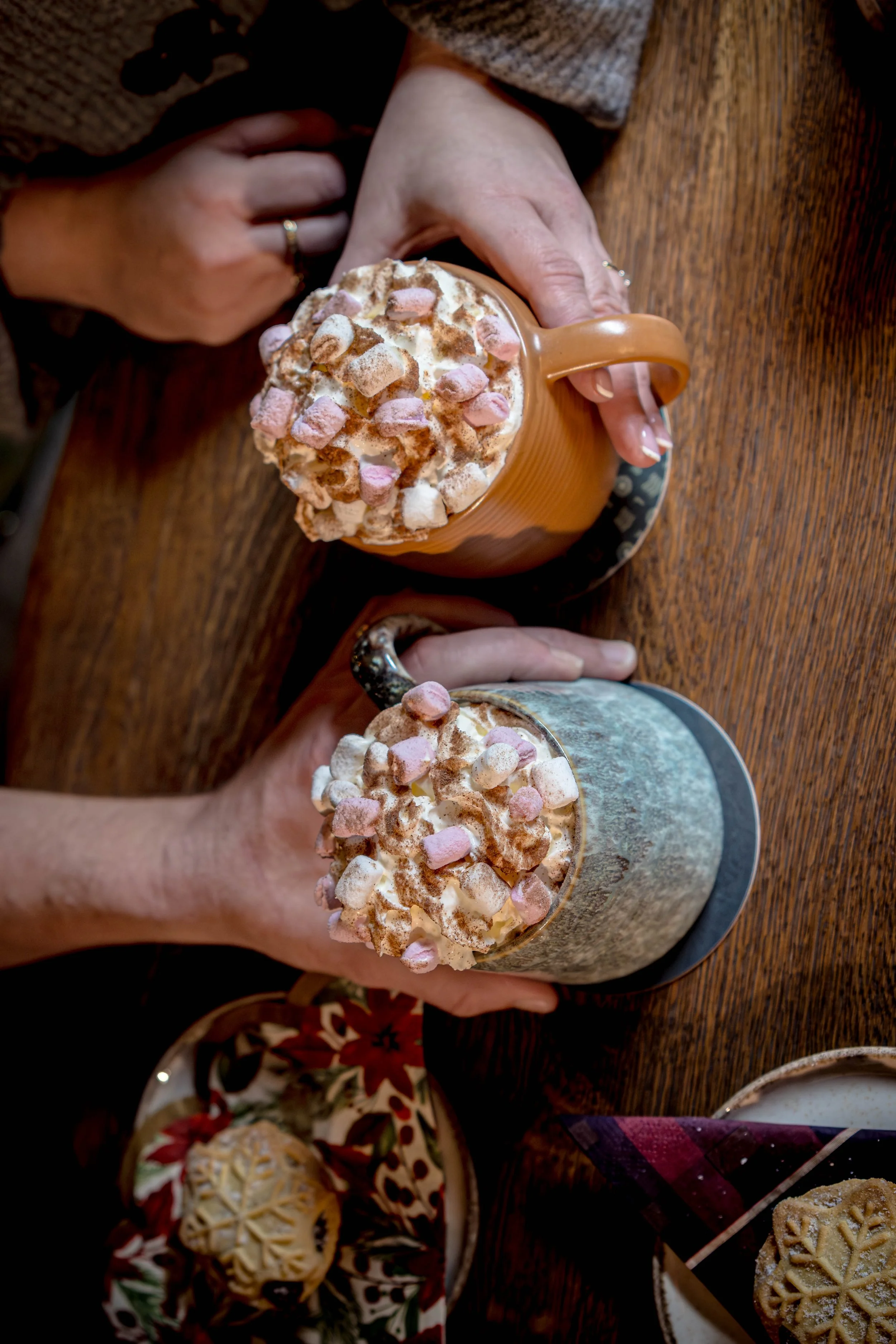 Two hands holding ceramic mugs topped with whipped cream, mini marshmallows, and cocoa powder, set on a wooden table with a plate of cookies nearby.