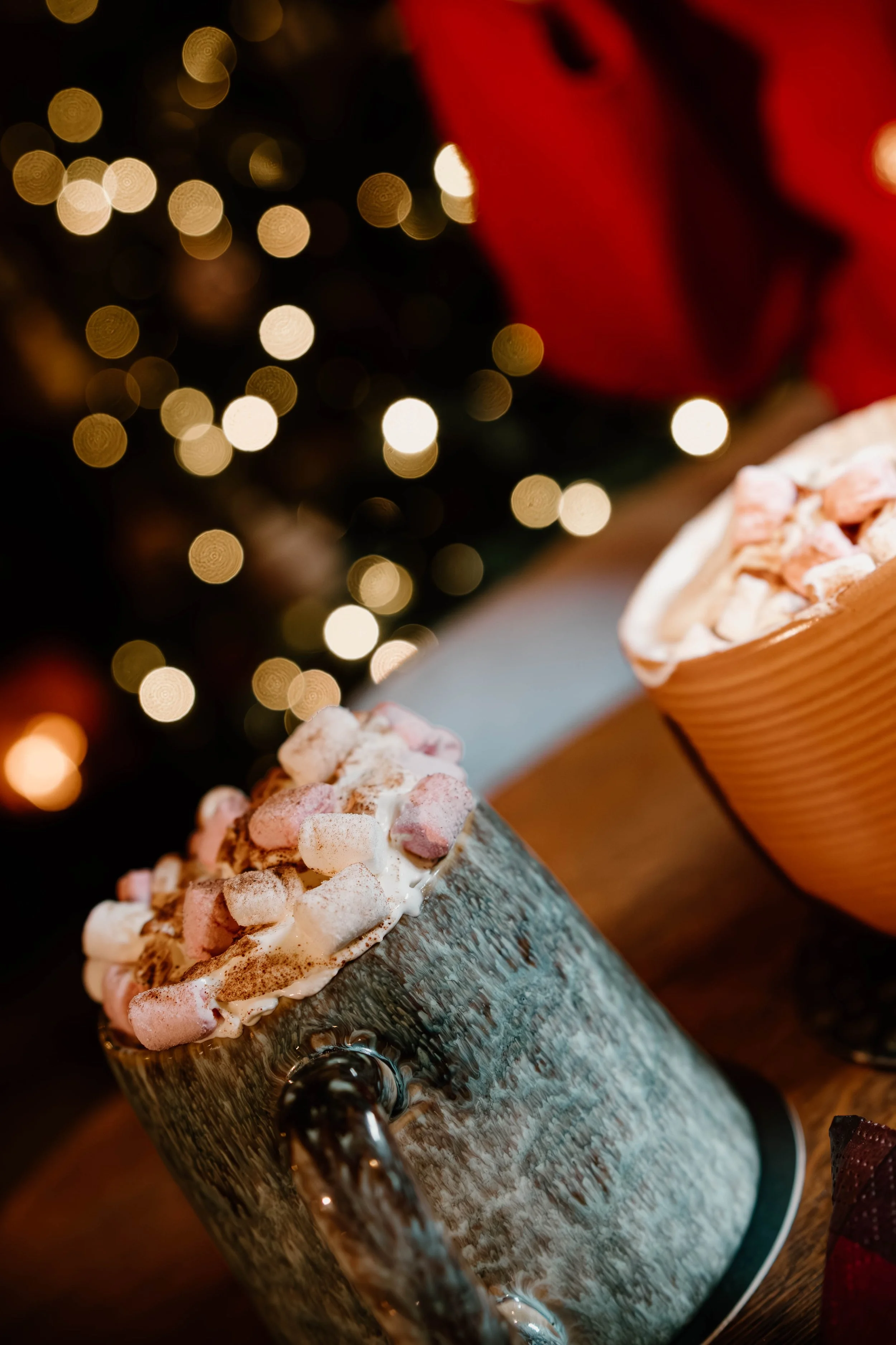 A mug filled with hot chocolate topped with mini marshmallows and whipped cream, set on a wooden surface with holiday lights in the background.