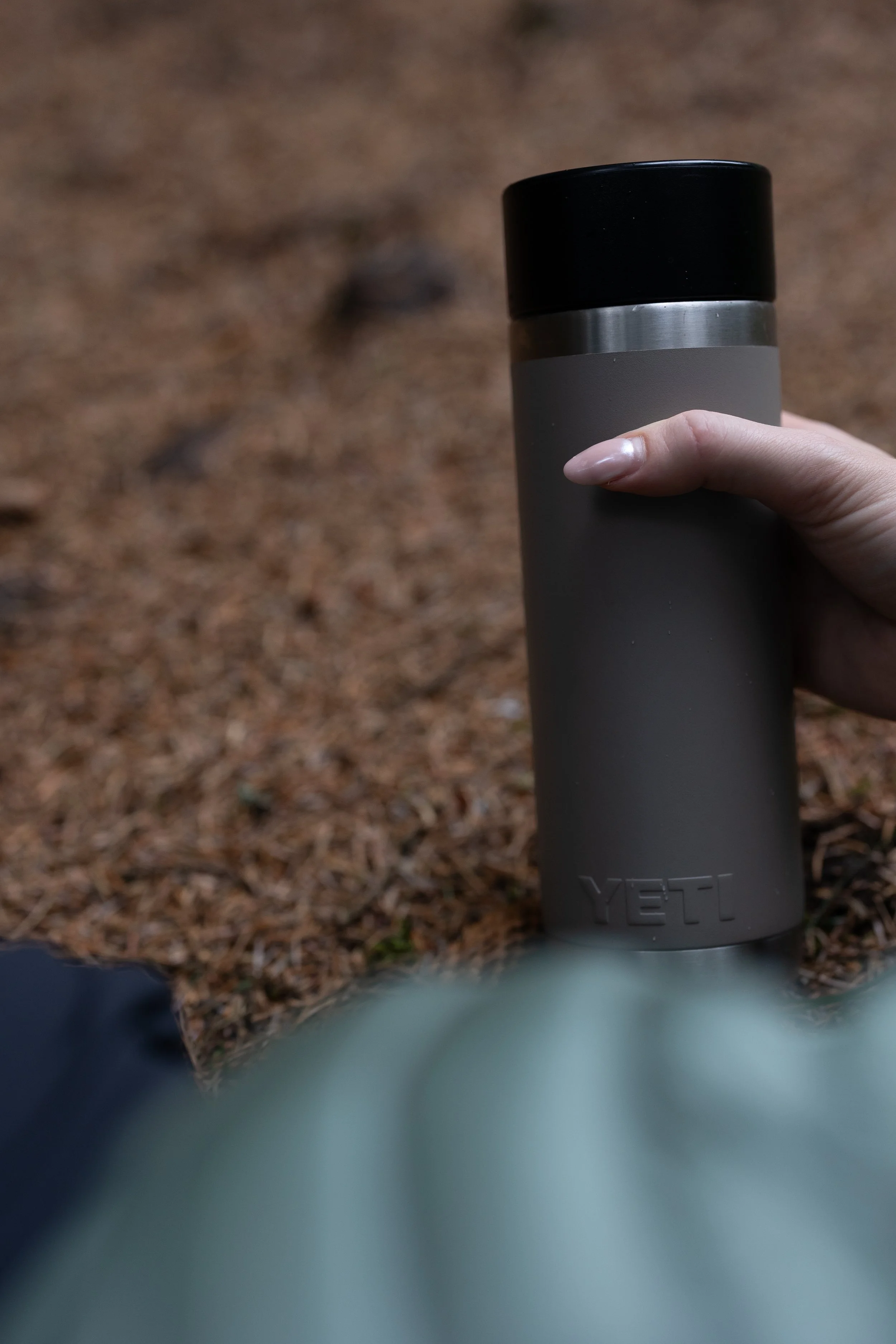 Close-up of a gray YETI thermos being held by a person outdoors on a ground covered with brown pine needles.