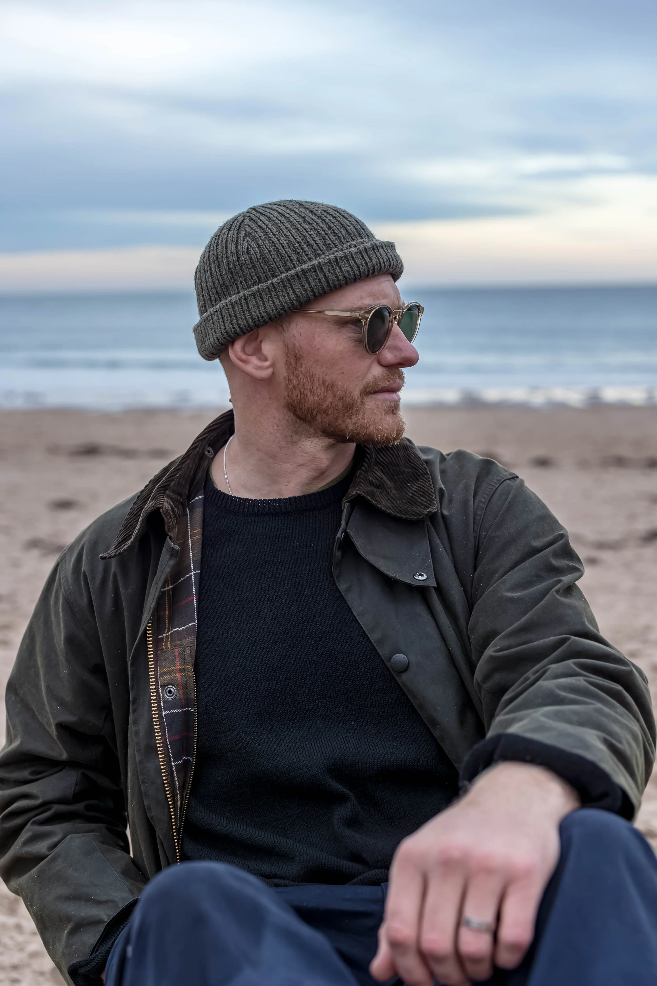 A man with red hair and beard sitting on a beach, wearing sunglasses, a knit cap, a black sweater, and a dark jacket, looking to the right with ocean in the background.