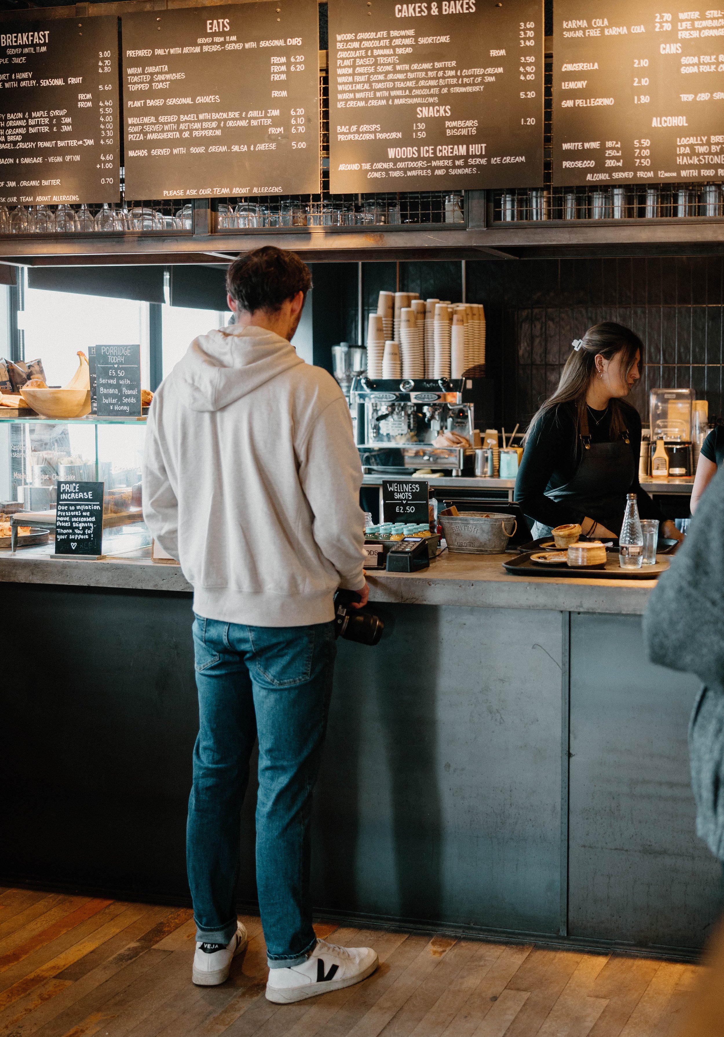 A man in a white hoodie and jeans stands at a cafe counter, holding a camera, as a female barista prepares drinks behind the counter.
