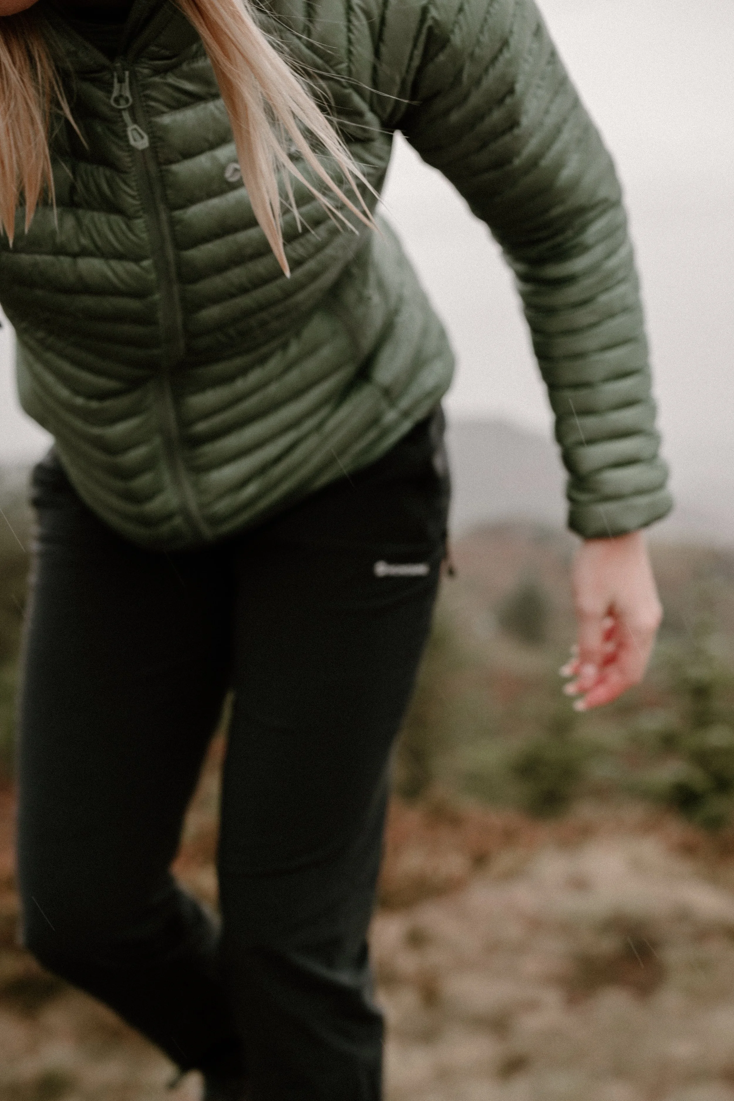 A person wearing a green puffer jacket and black pants, standing outdoors on a foggy day.