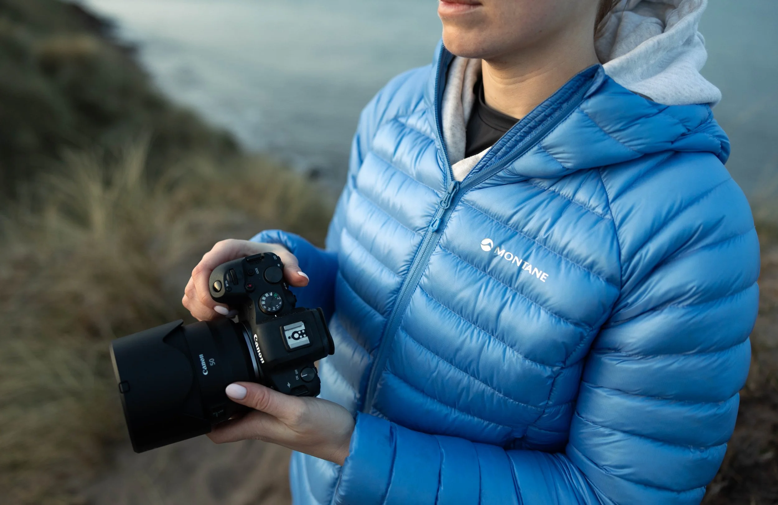 Person in a blue Montane jacket holding a Canon camera outdoors near water, with grassy terrain in the background.