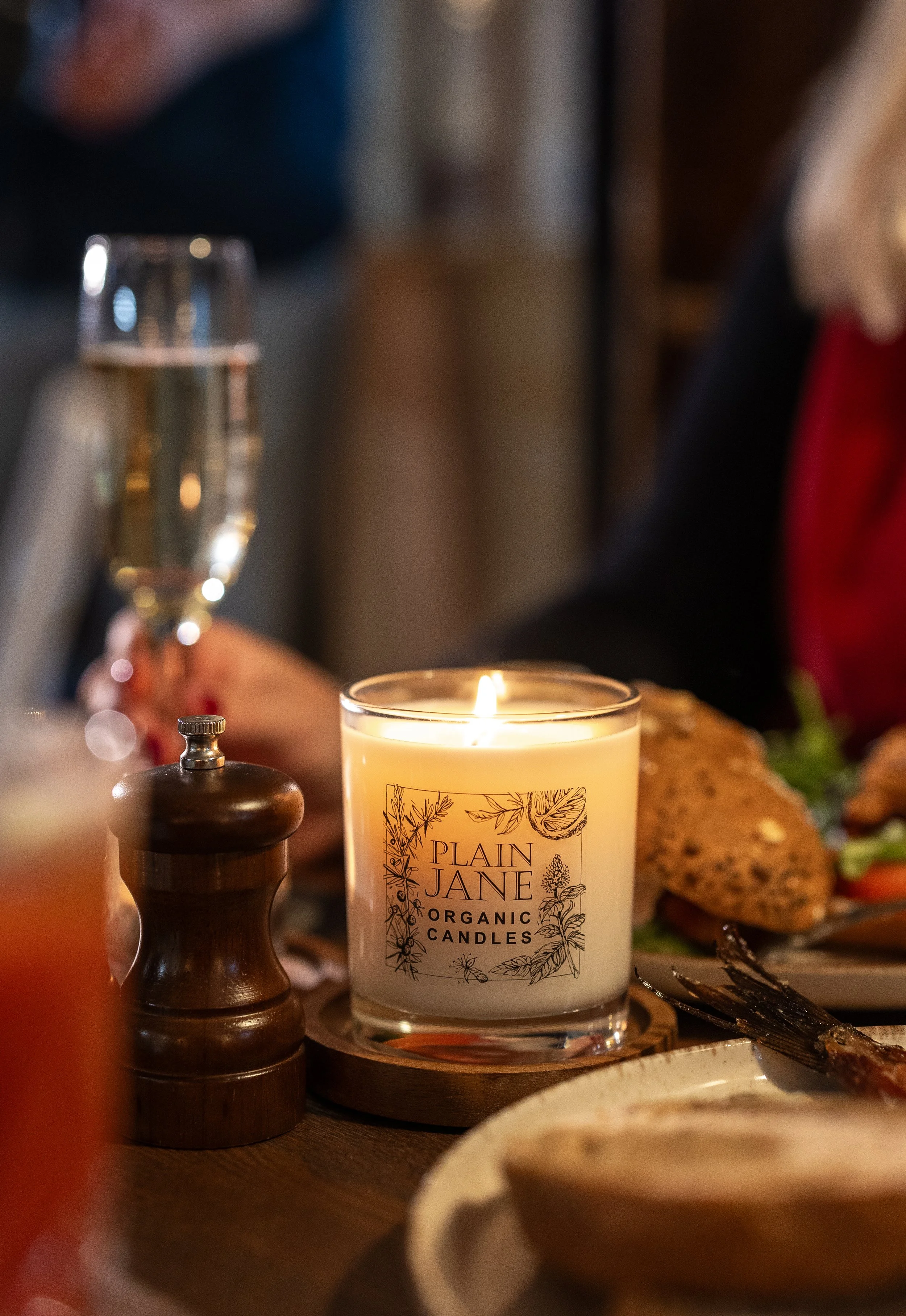 A lit candle labeled 'Plain Jane Organic Candles' on a wooden table, with a glass of champagne, a pepper grinder, and a plate of food in the background.