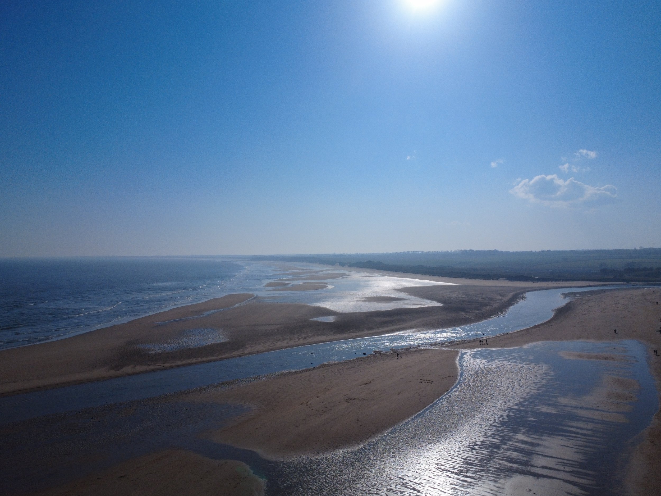A wide view of a beach with sand dunes, small streams, and the ocean under a partly cloudy sky.