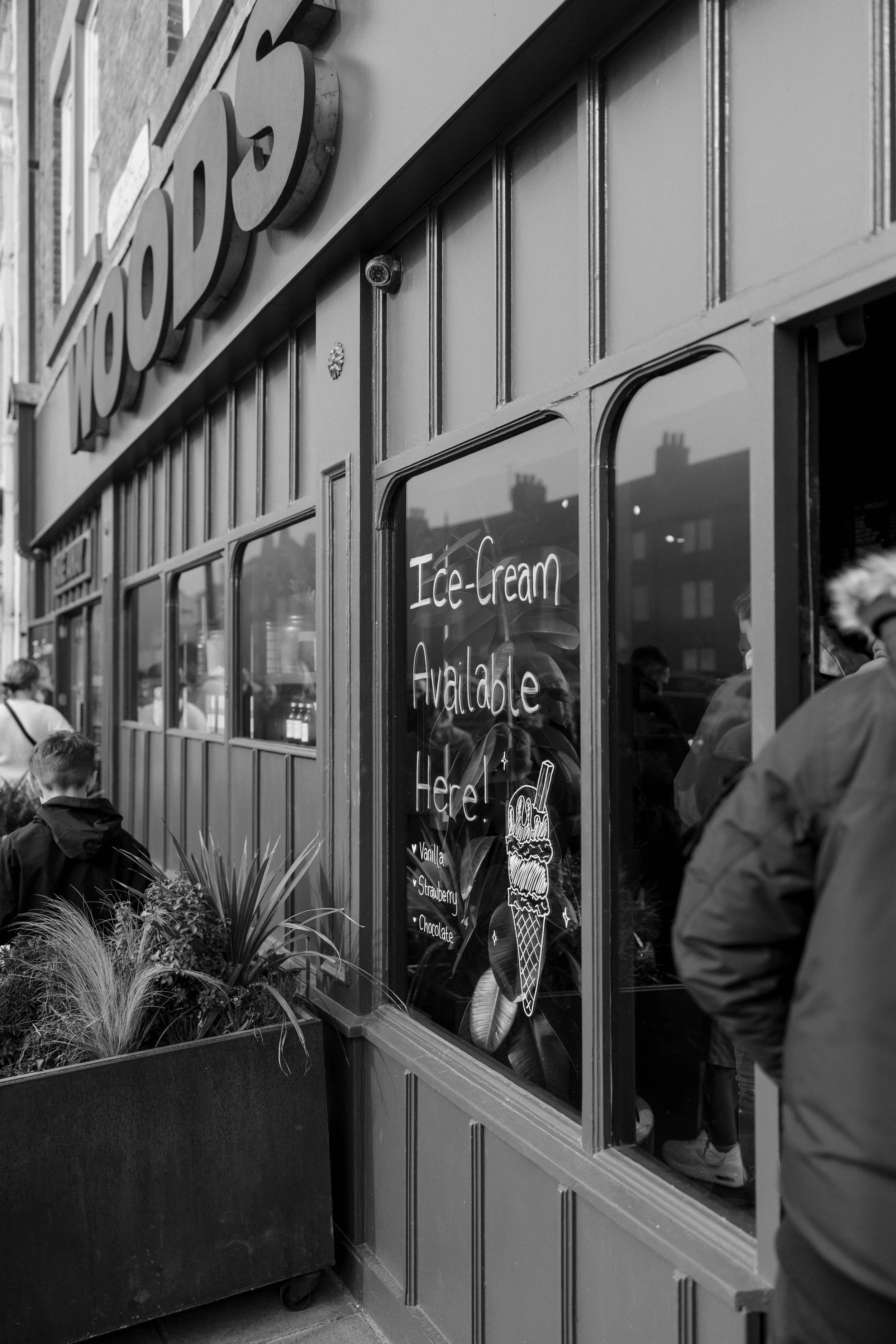 Black and white photo of an ice cream shop with a sign on the window that reads "Ice-Cream Available Here!" and lists flavors: vanilla, strawberry, and chocolate. People are seen outside and inside the shop, along with potted plants outside the windo