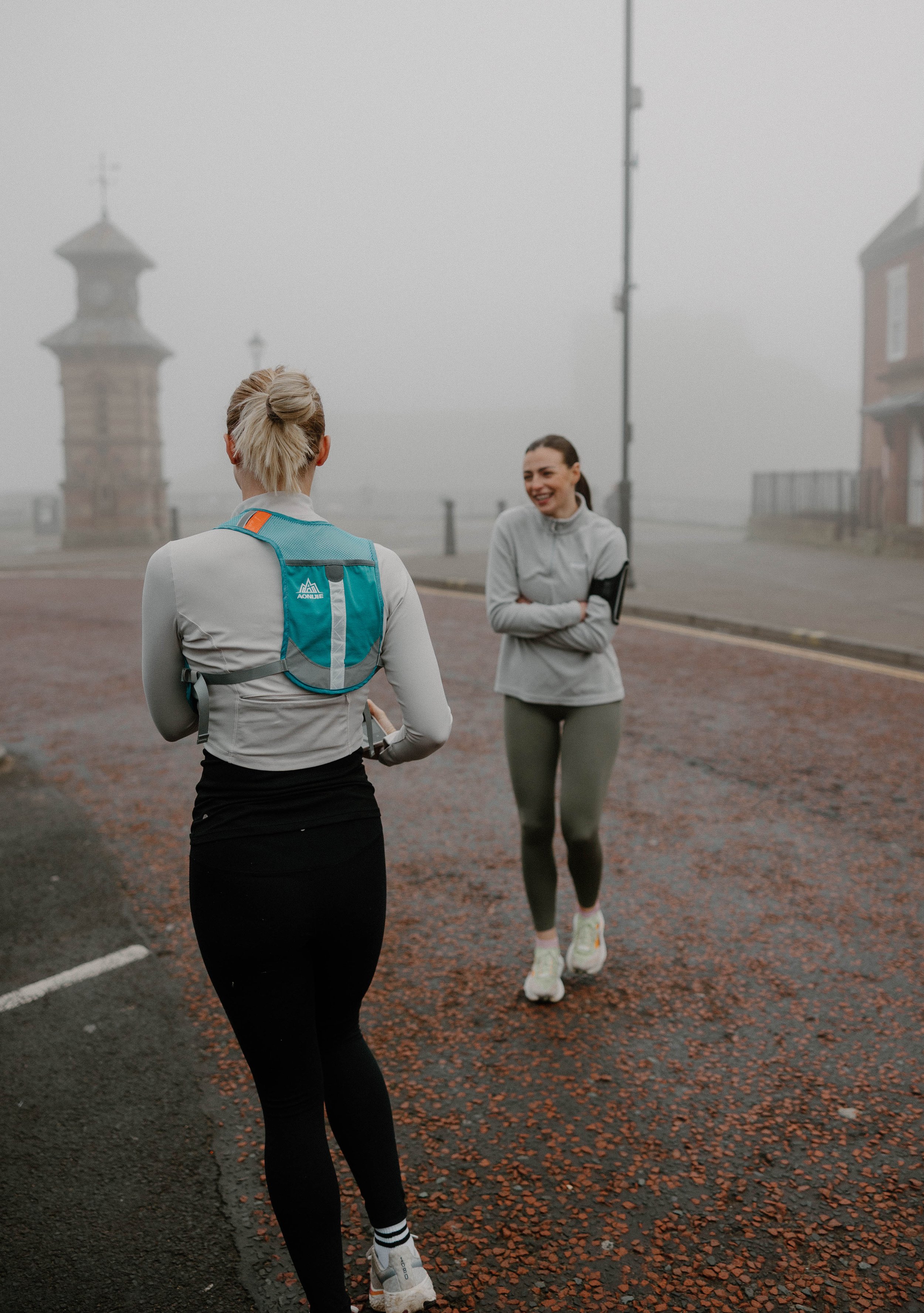 Two women in athletic clothing and running shoes talking outdoors on a foggy day, with one facing away and the other smiling.