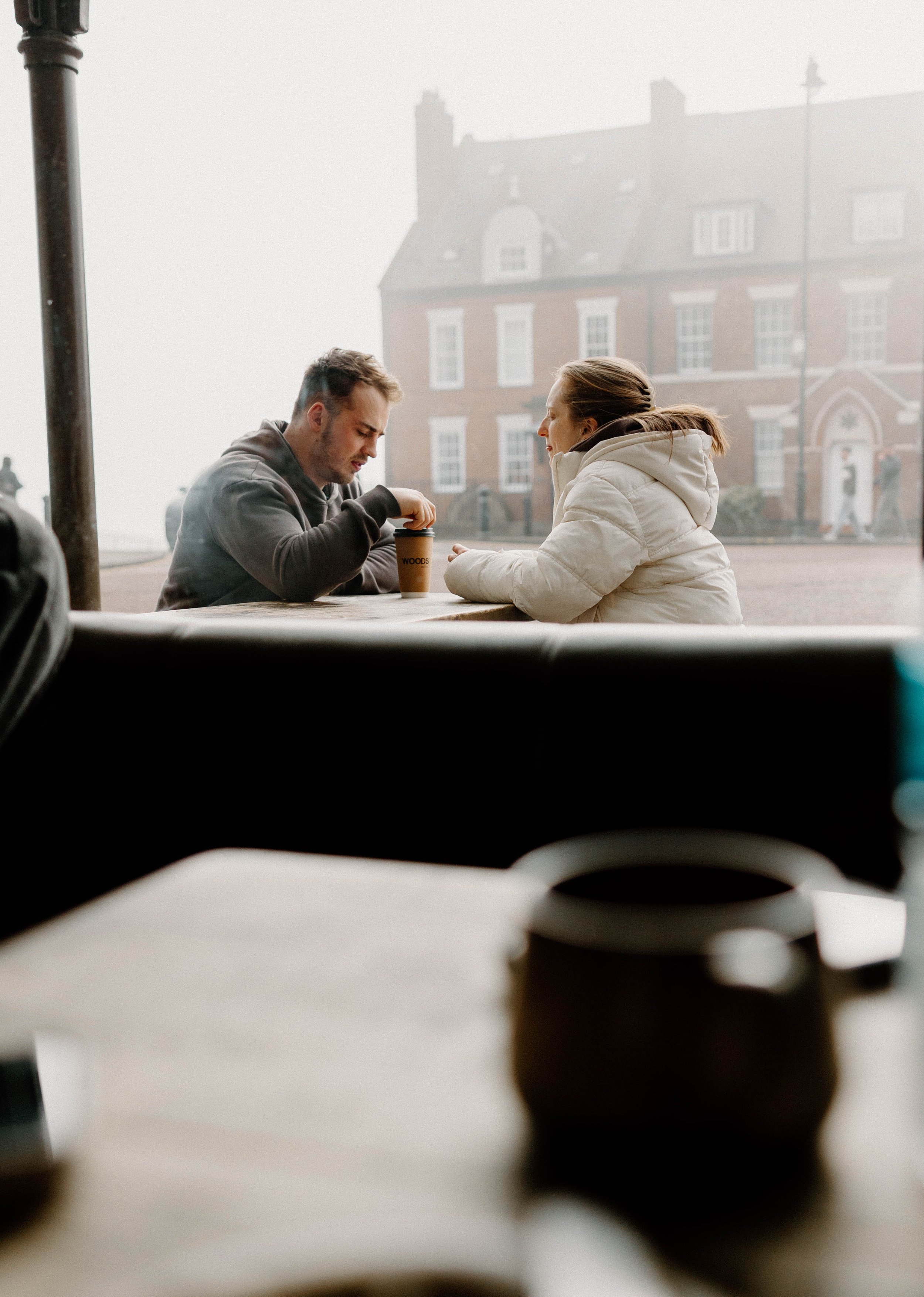 Two people sitting at an outdoor table at a café, with a city street and old brick building in the background, foggy weather.