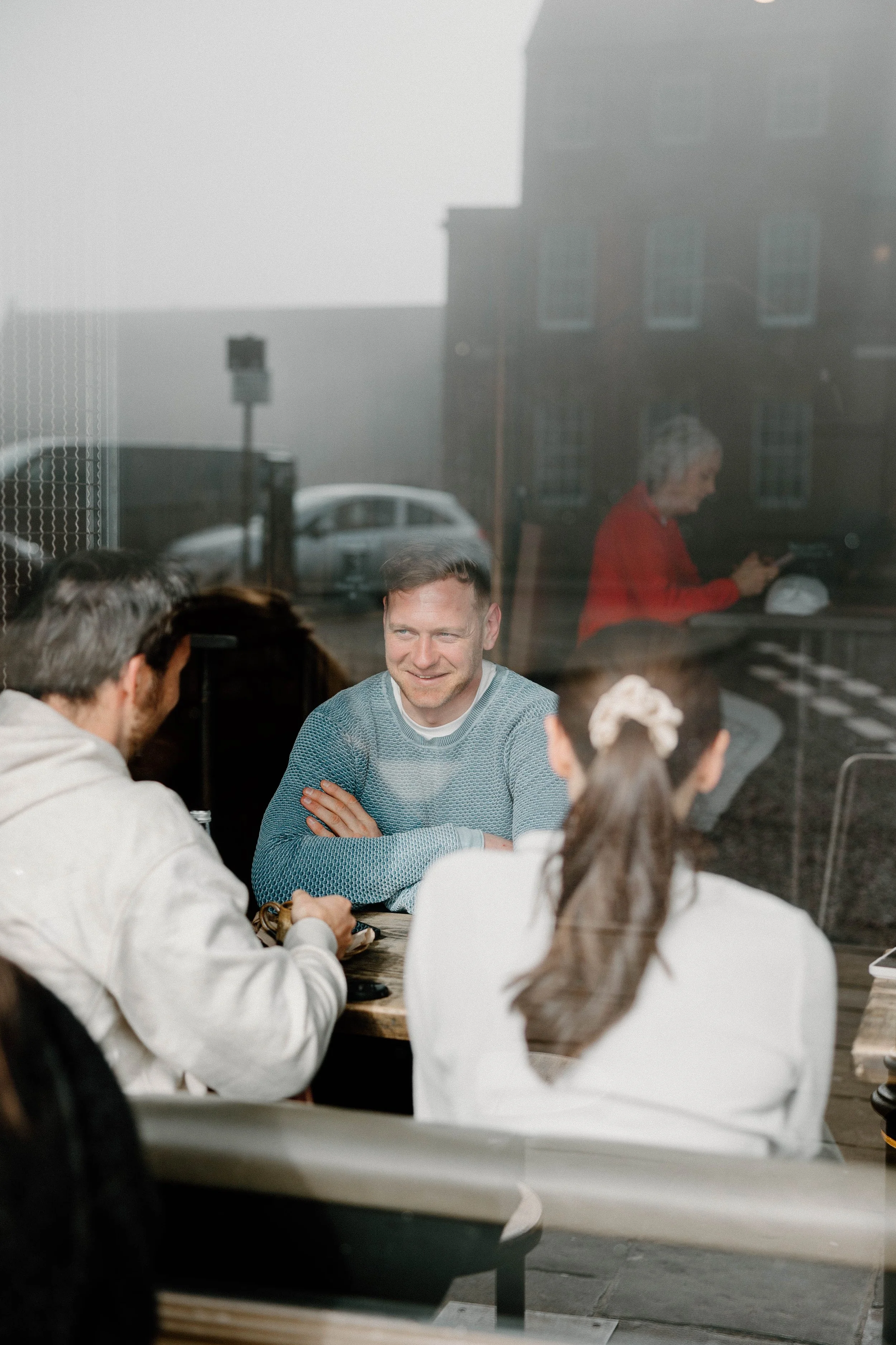 People sitting at an outdoor cafe table, seen through a large window, with a man smiling at others in the group and a woman in a white top with a hair accessory