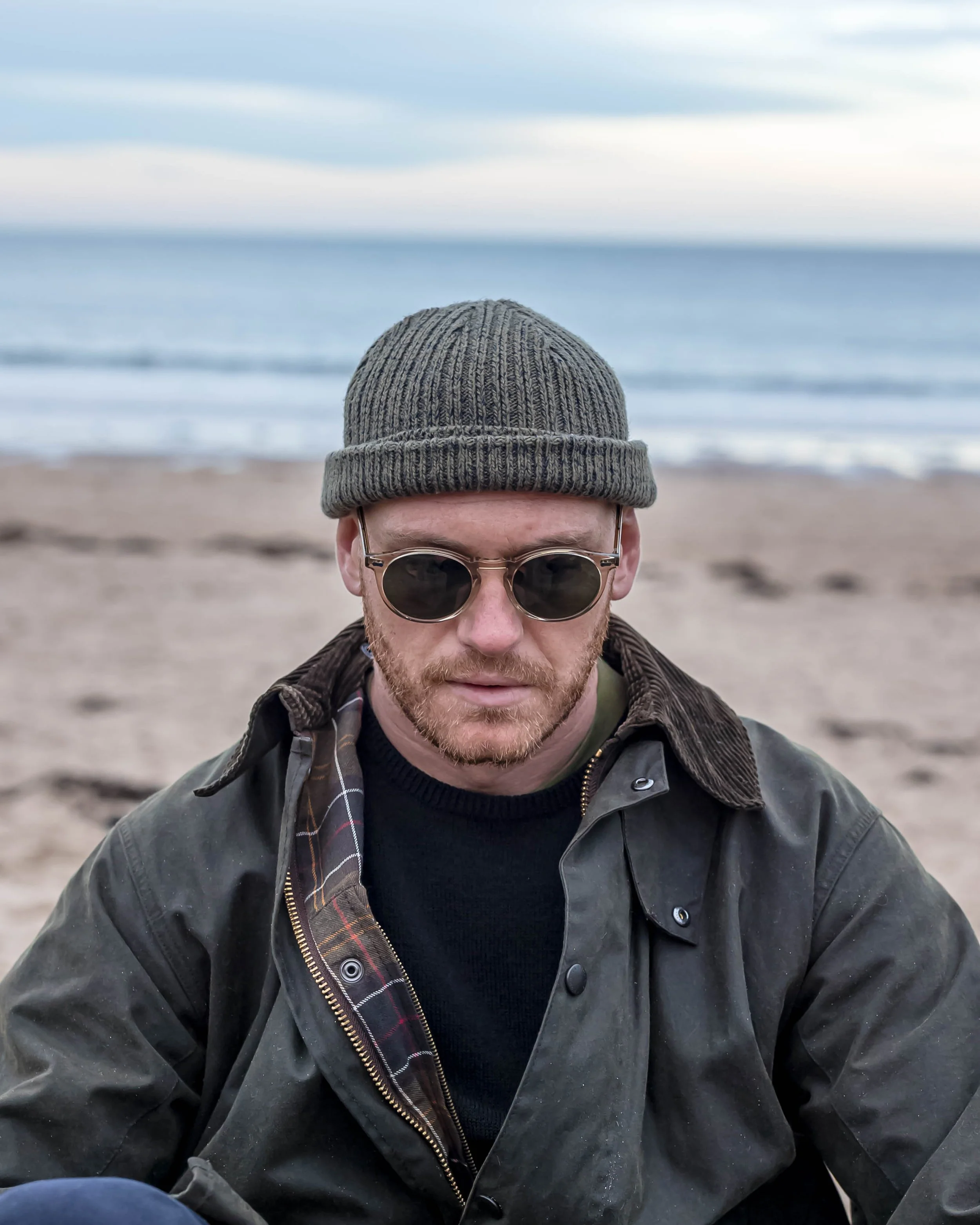 A man in a gray knit beanie, sunglasses, black jacket, and plaid shirt, sitting on a beach with the ocean and cloudy sky in the background.