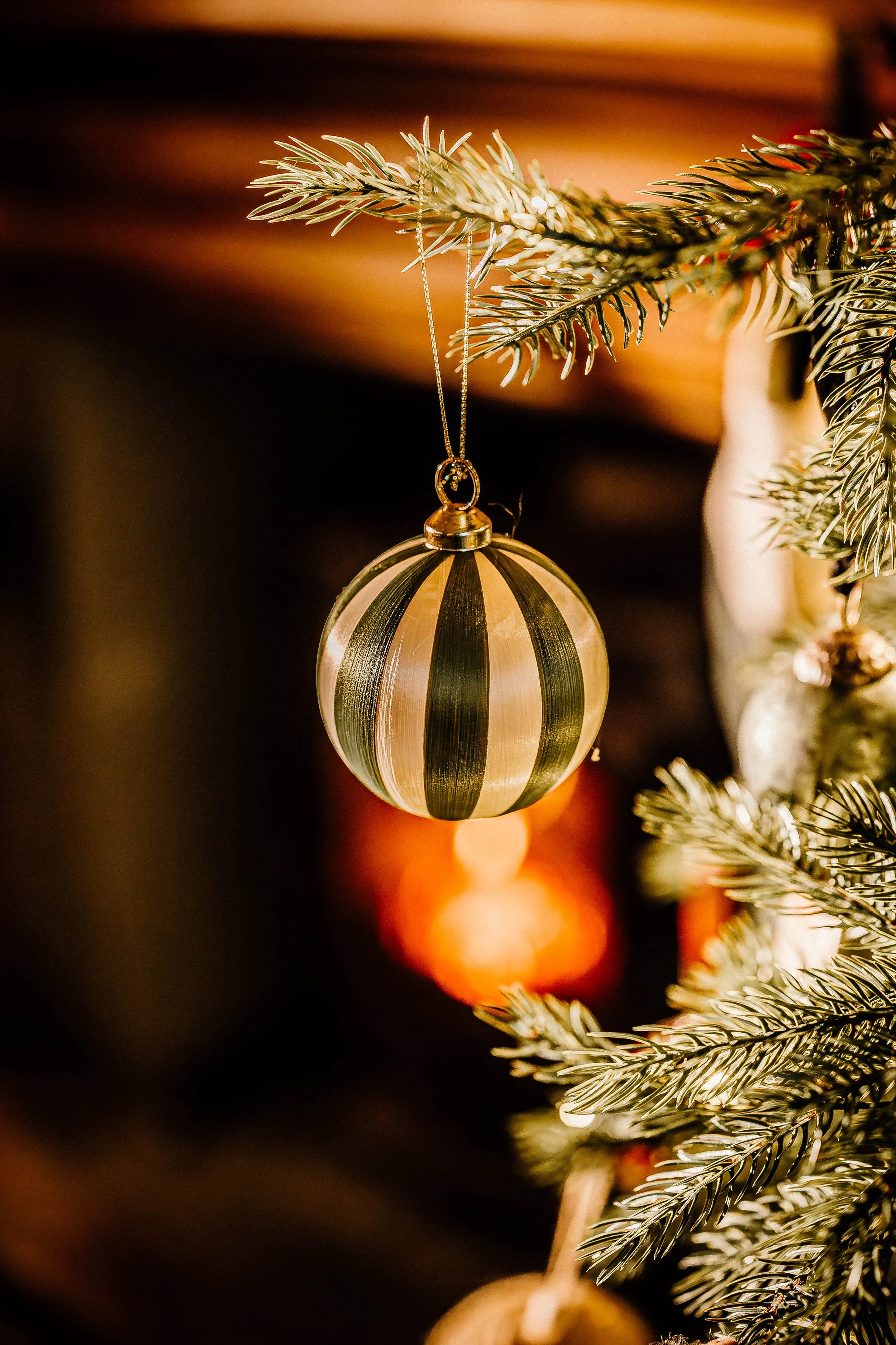 A close-up of a Christmas tree ornament hanging from a branch of a decorated Christmas tree. The ornament is round, gold and black striped, with a shiny surface, and it is suspended by a thin gold string.