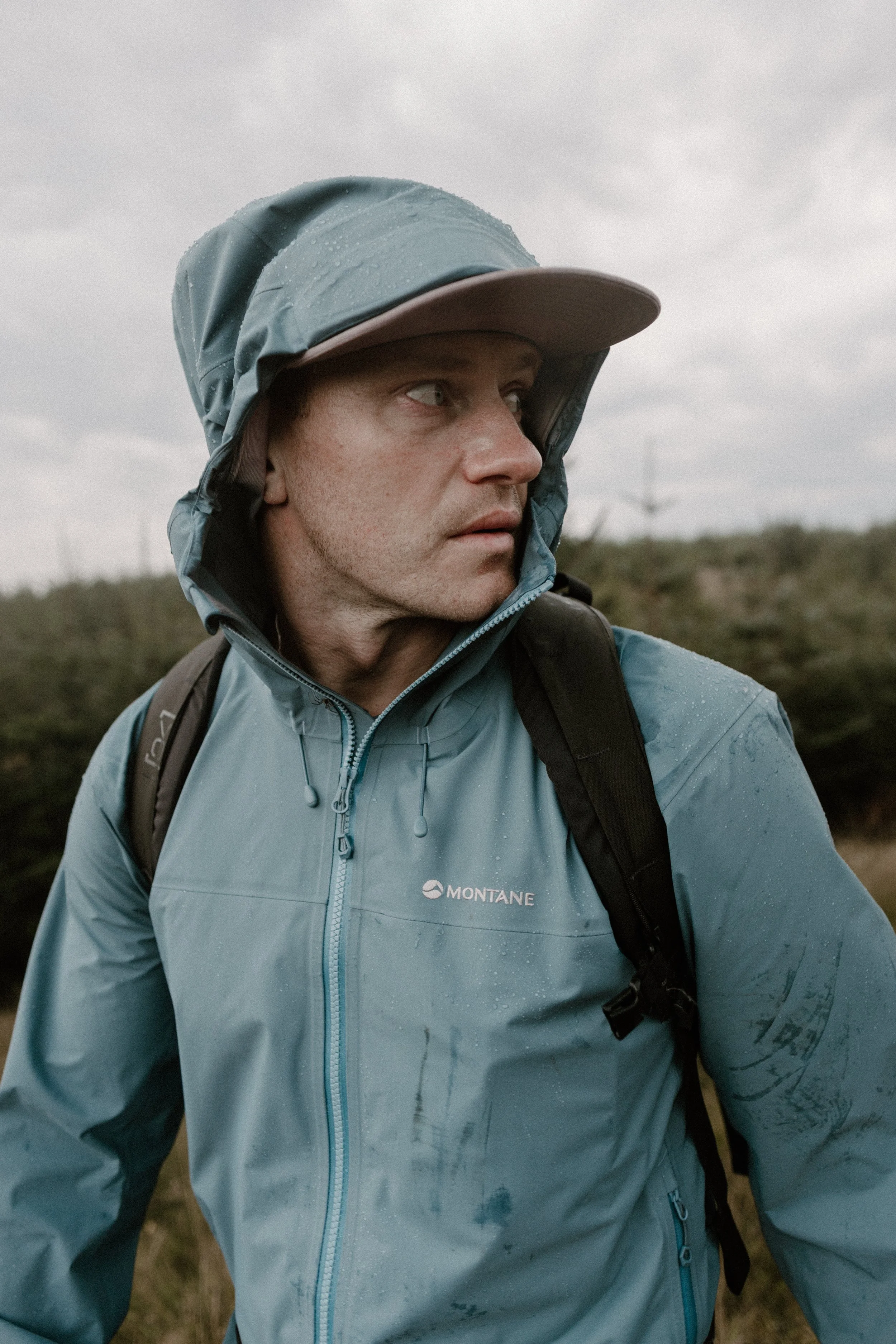 A man in rain gear outdoors, wearing a light blue waterproof jacket with droplets of rain on it, a hat, and a backpack, looking to his left against a cloudy sky and greenery.