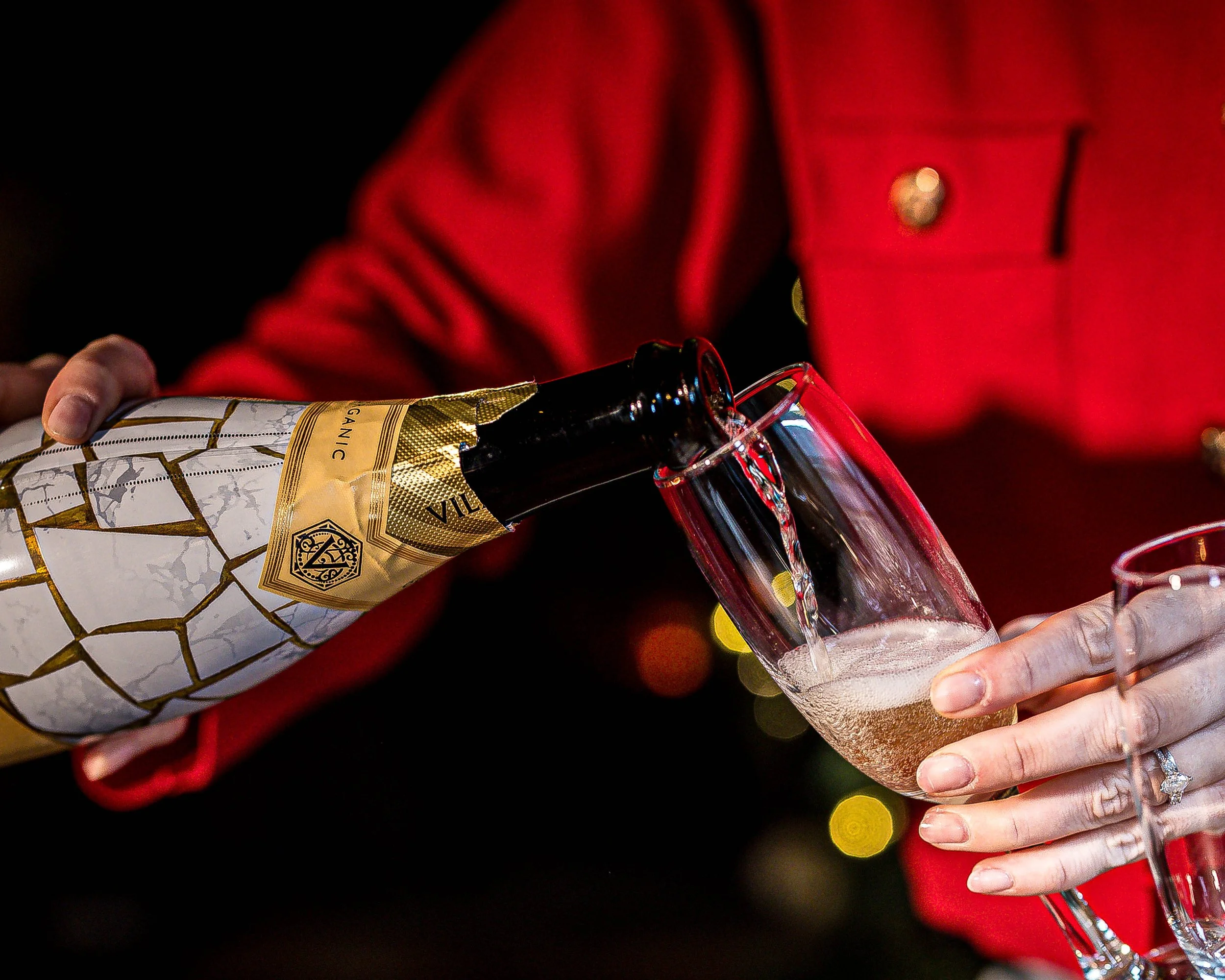 Person pouring champagne into a glass, wearing a red jacket.