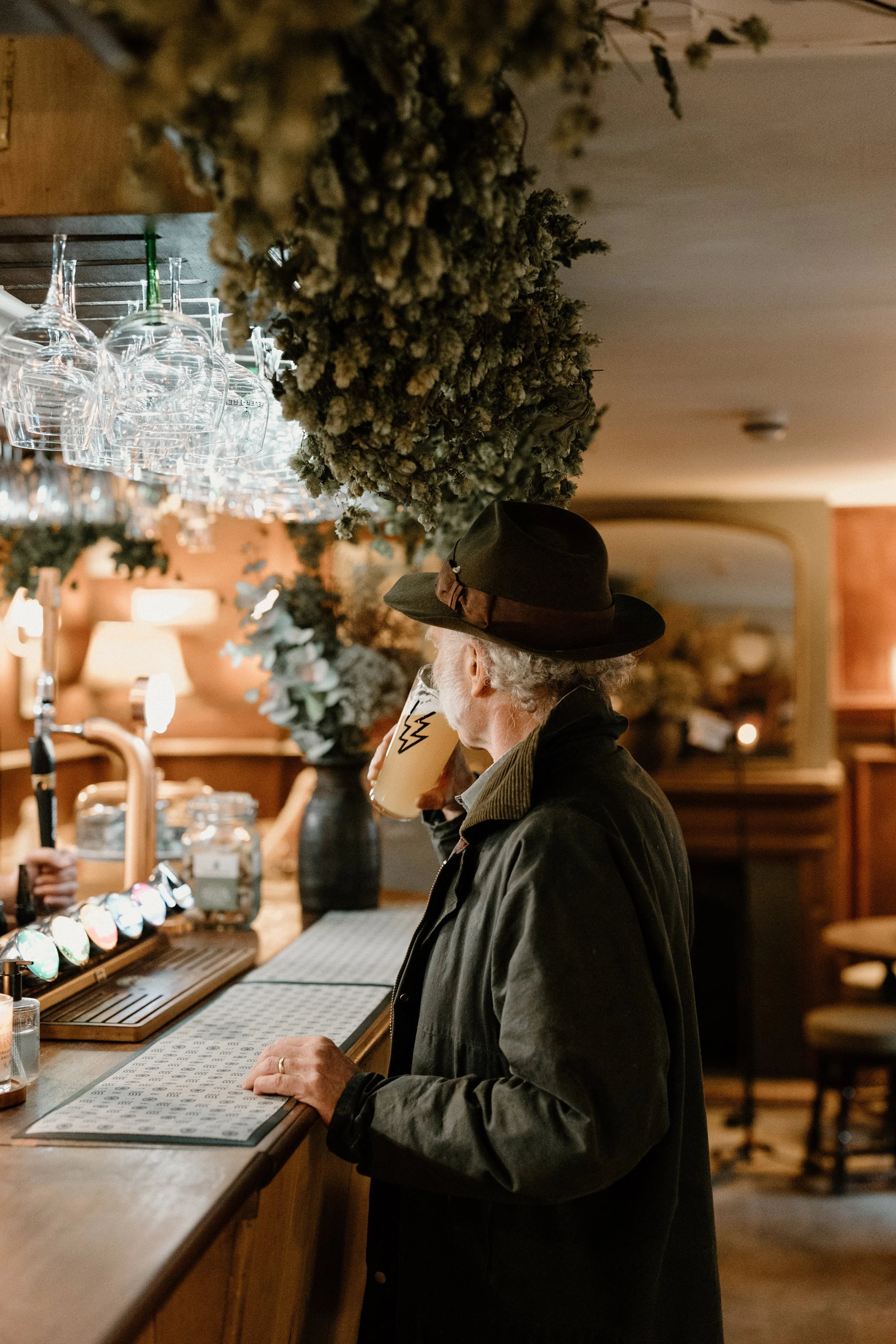 An elderly man with gray hair, wearing a dark hat and jacket, is drinking a beer at a bar counter decorated with dried herbs and flowers, inside a cozy pub or bar.