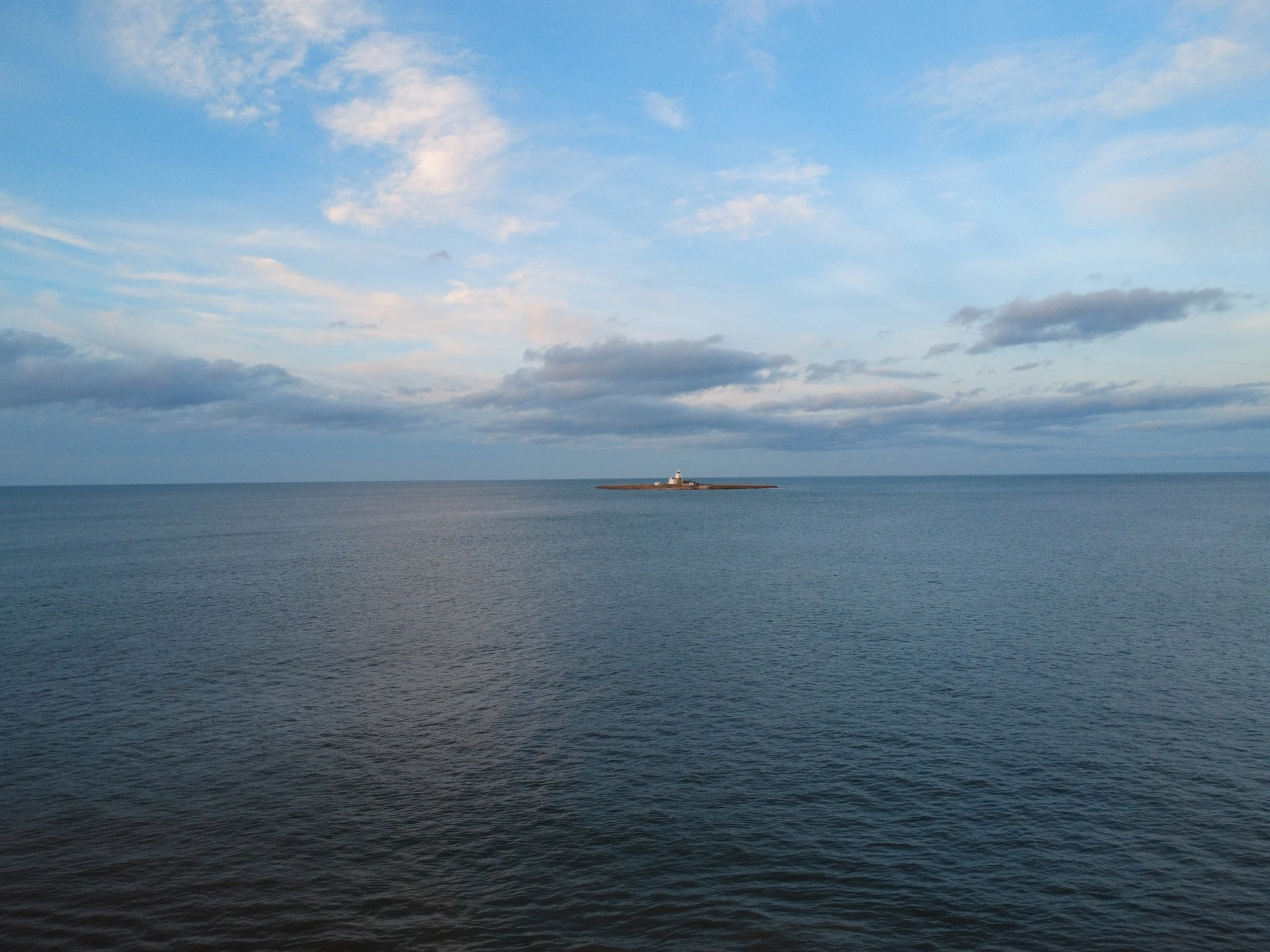 Ocean with a small lighthouse on a rocky outcrop in the distance under a partly cloudy sky.