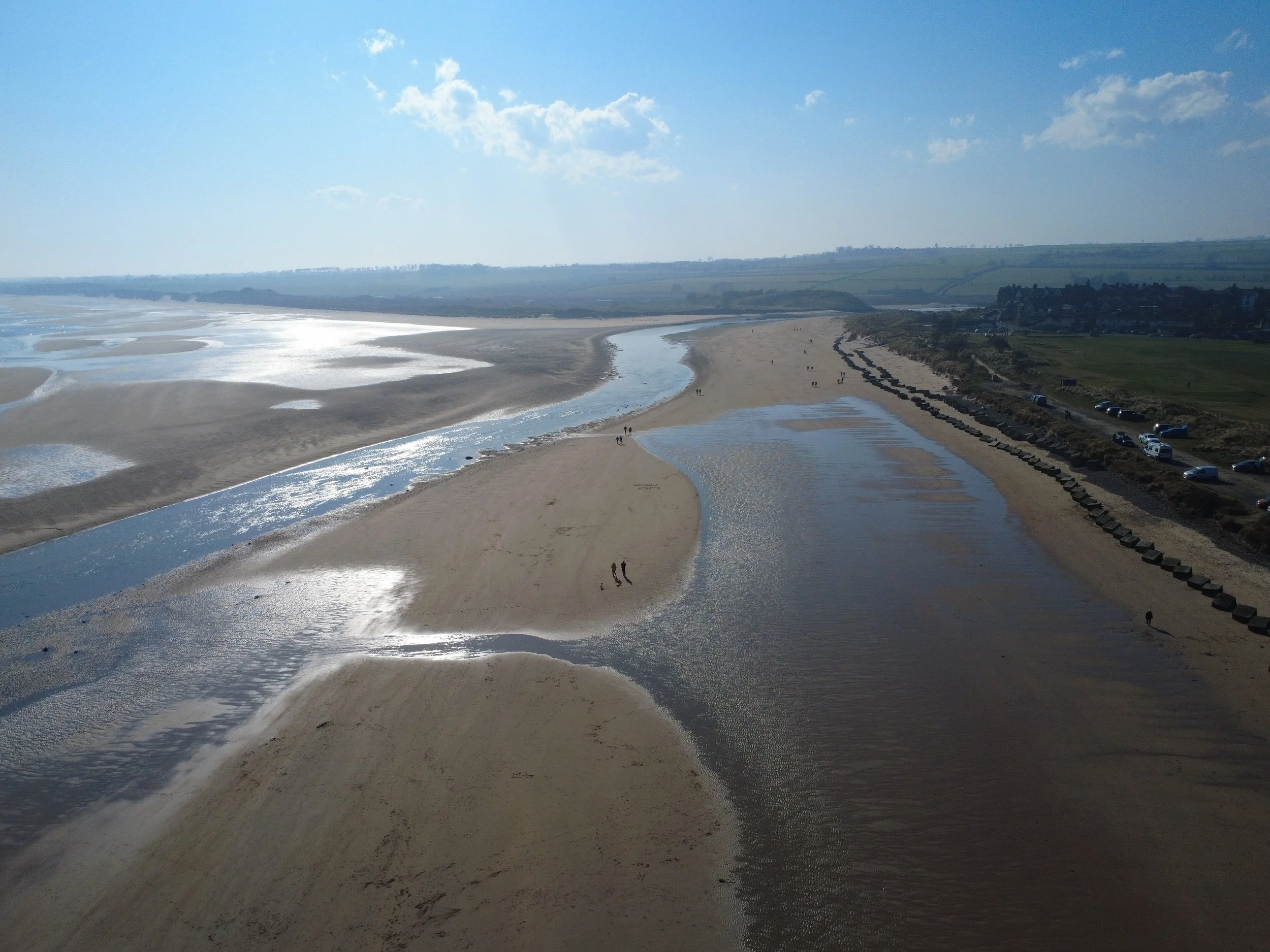 A beach scene with a sandy shoreline, shallow tidal pools, and a river flowing into the sea under a partly cloudy sky.