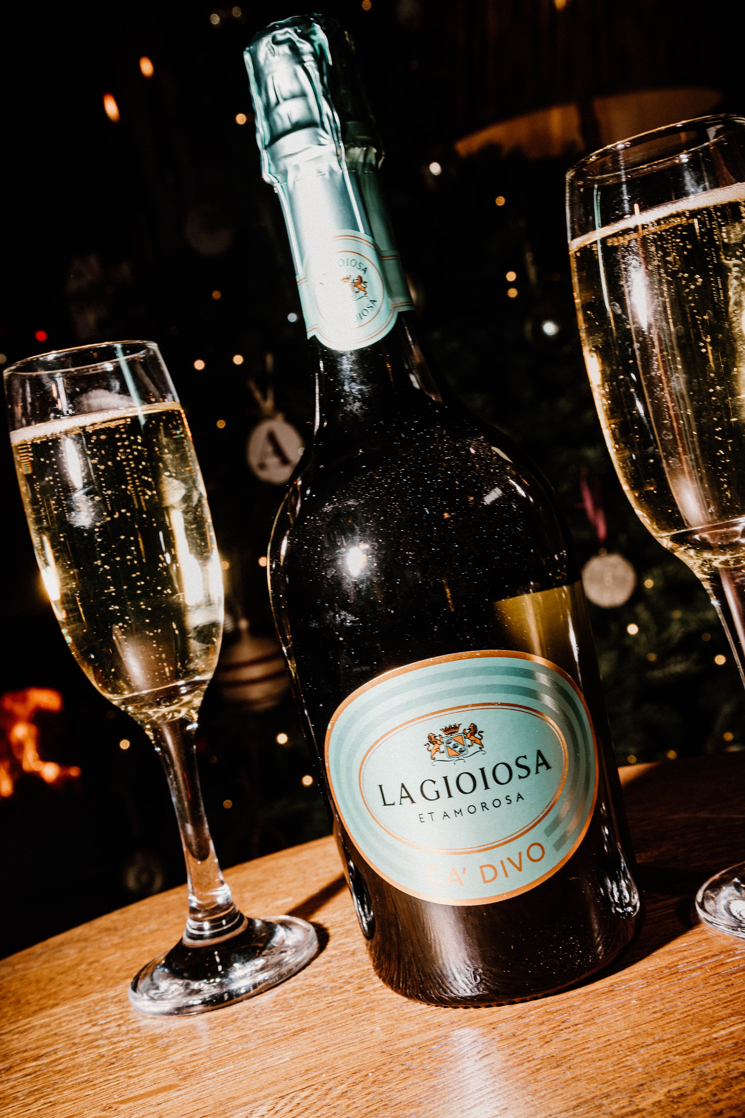A bottle of Lagioisa Etamorosa sparkling wine surrounded by two filled champagne glasses on a wooden table with a blurred festive background.