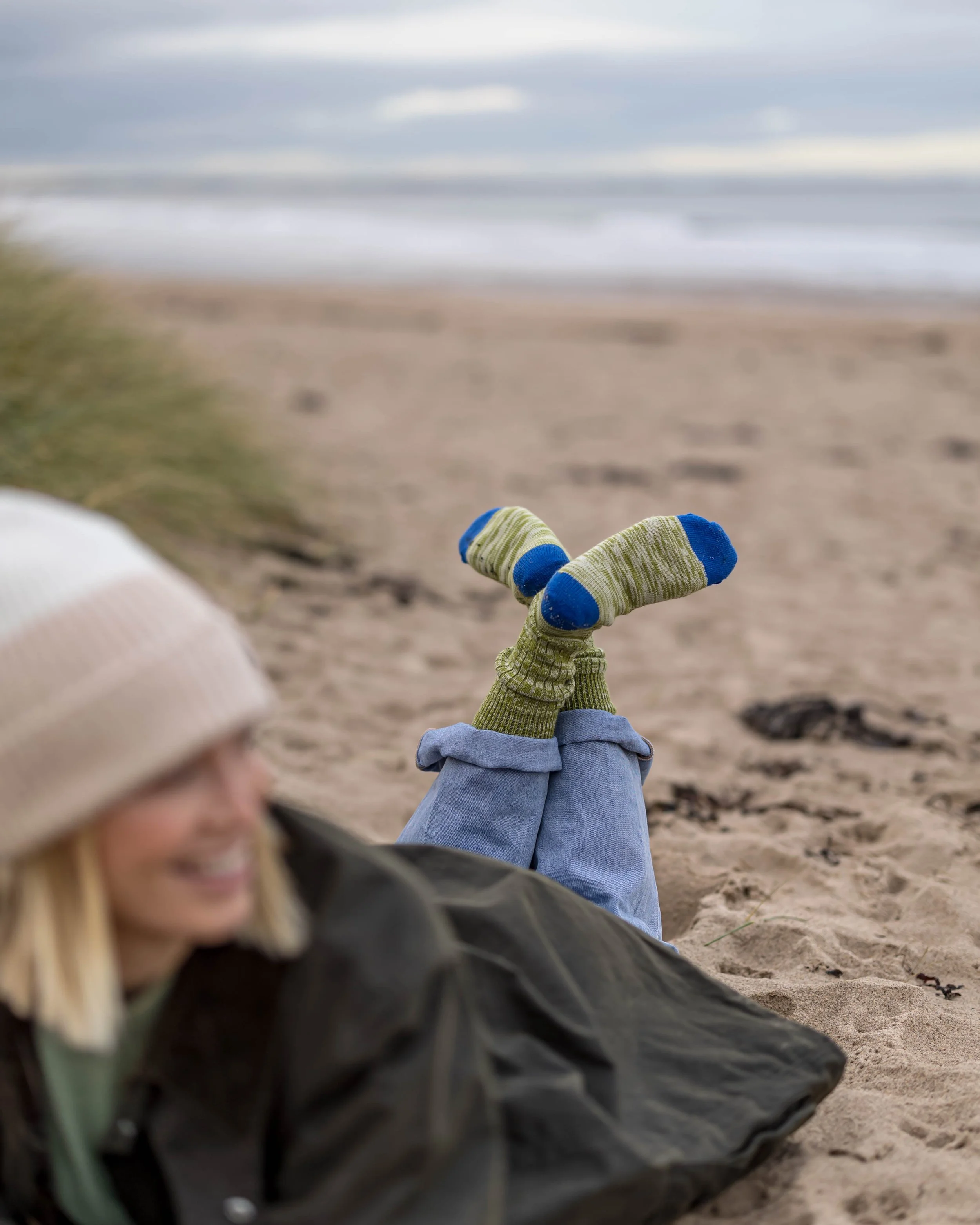 Person lying on the sand with feet raised, wearing striped socks and jeans, near the beach with waves in the distance, under cloudy sky.