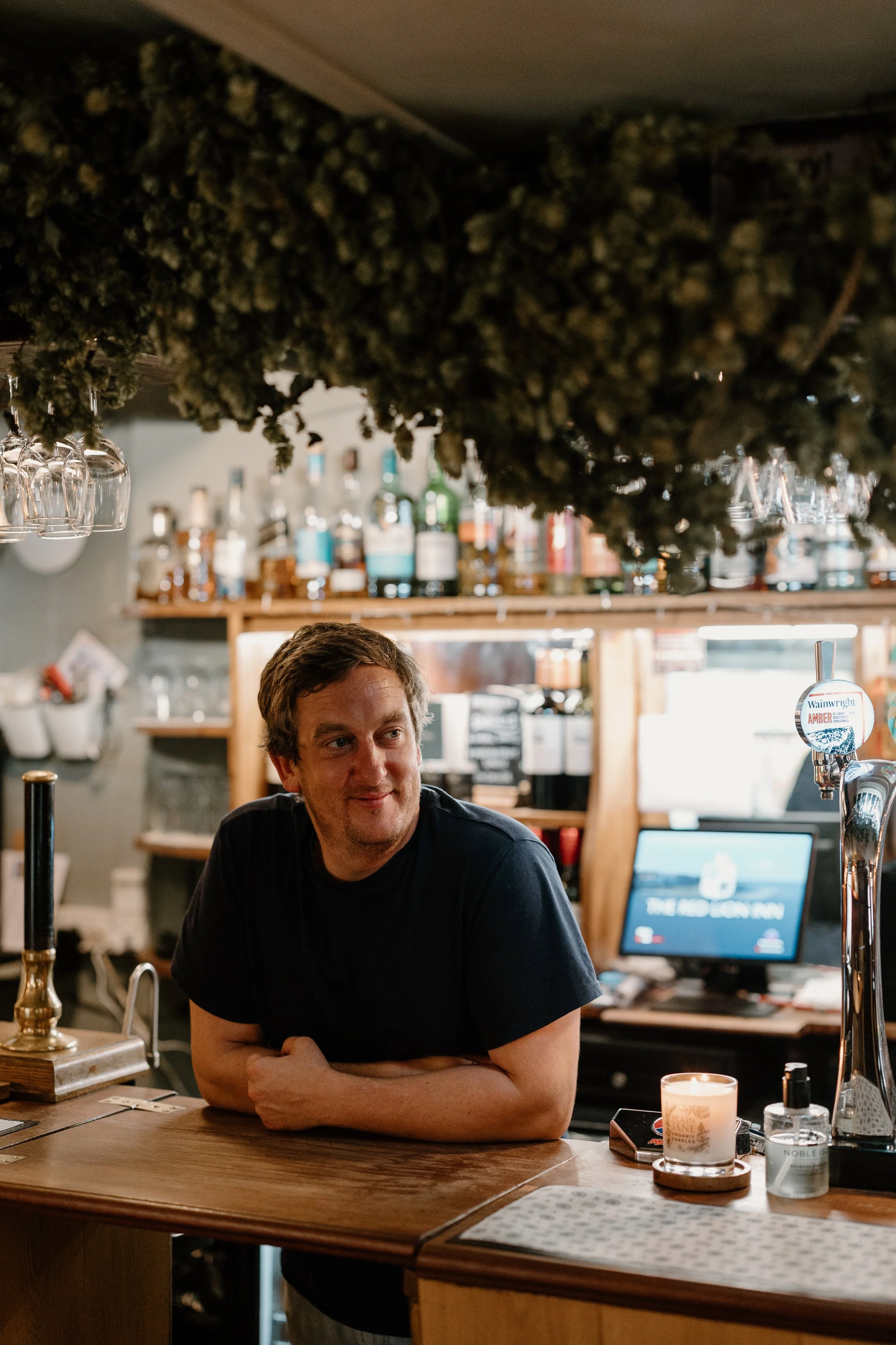 A man with brown hair and a beard leaning on a wooden bar counter with a smile, in a bar setting with bottles on shelves, glassware, and a computer in the background, and a lit candle on the counter.