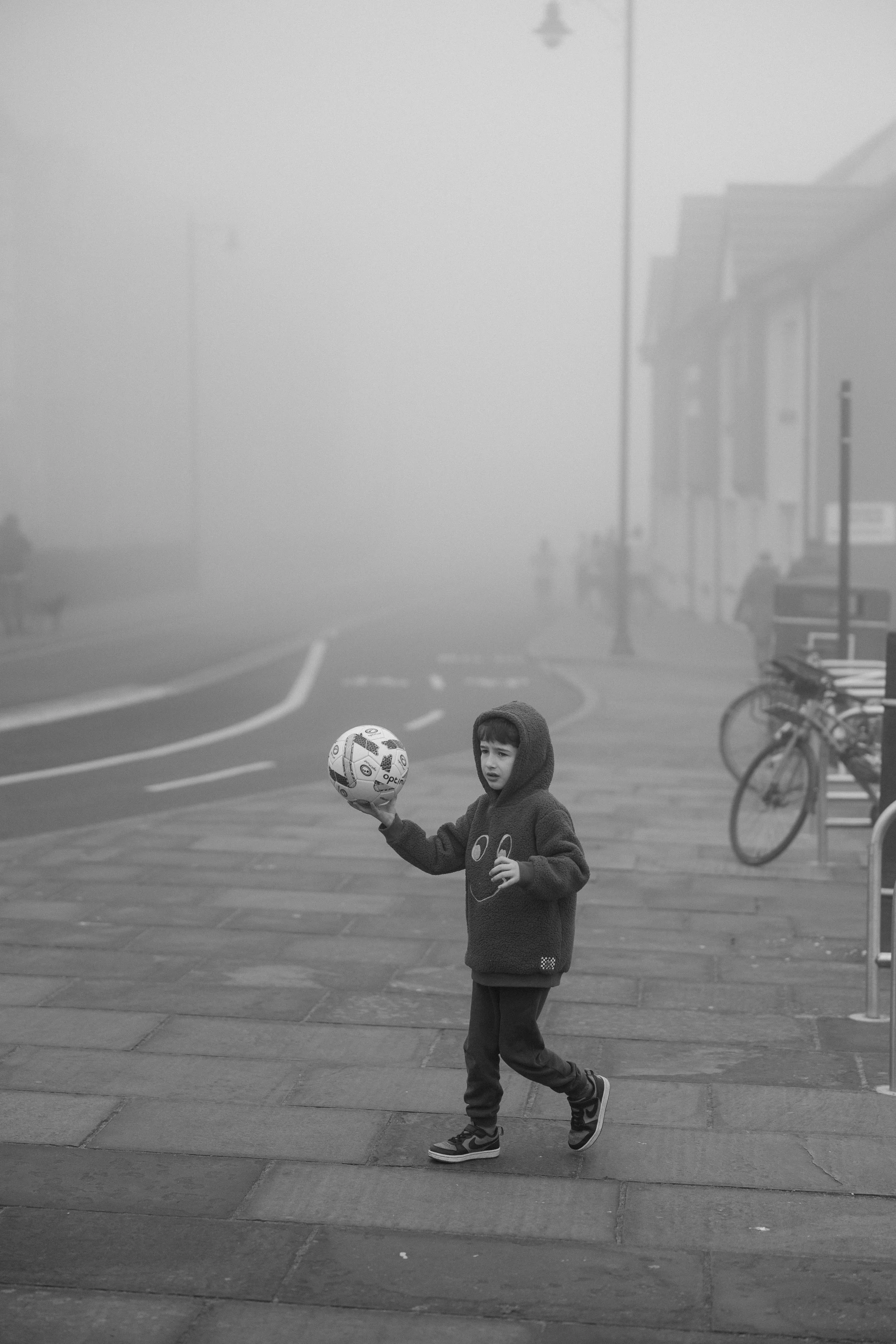 A young boy wearing a hoodie and sweatpants is holding a soccer ball on a foggy street sidewalk with bicycles parked nearby.