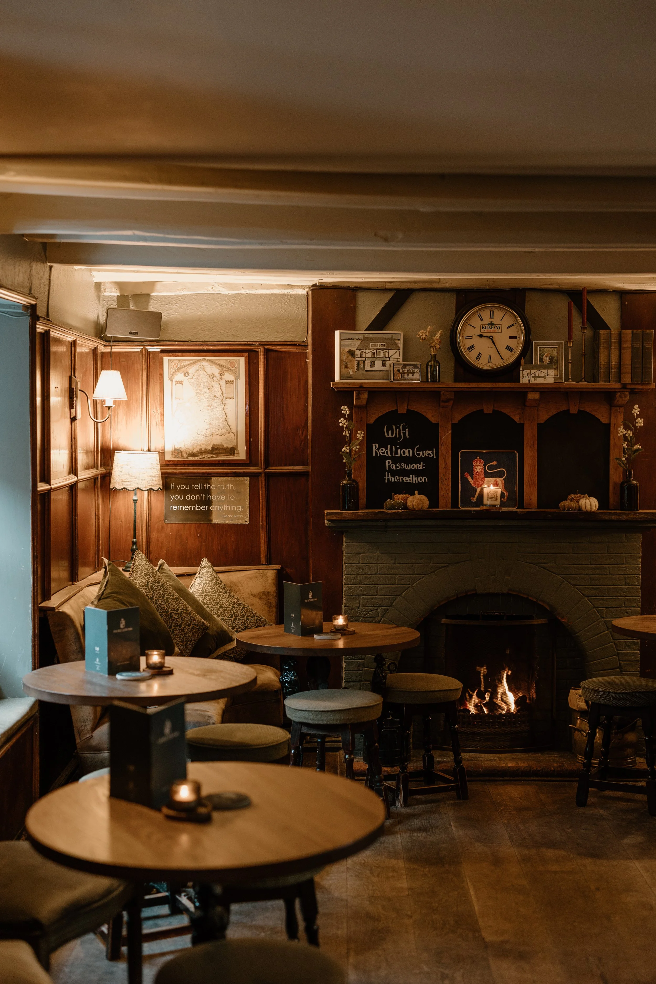 Cozy restaurant with a brick fireplace, wooden tables, upholstered chairs, wall decor, and a chalkboard with Wi-Fi and password details. Dim lighting and autumn-themed decorations.