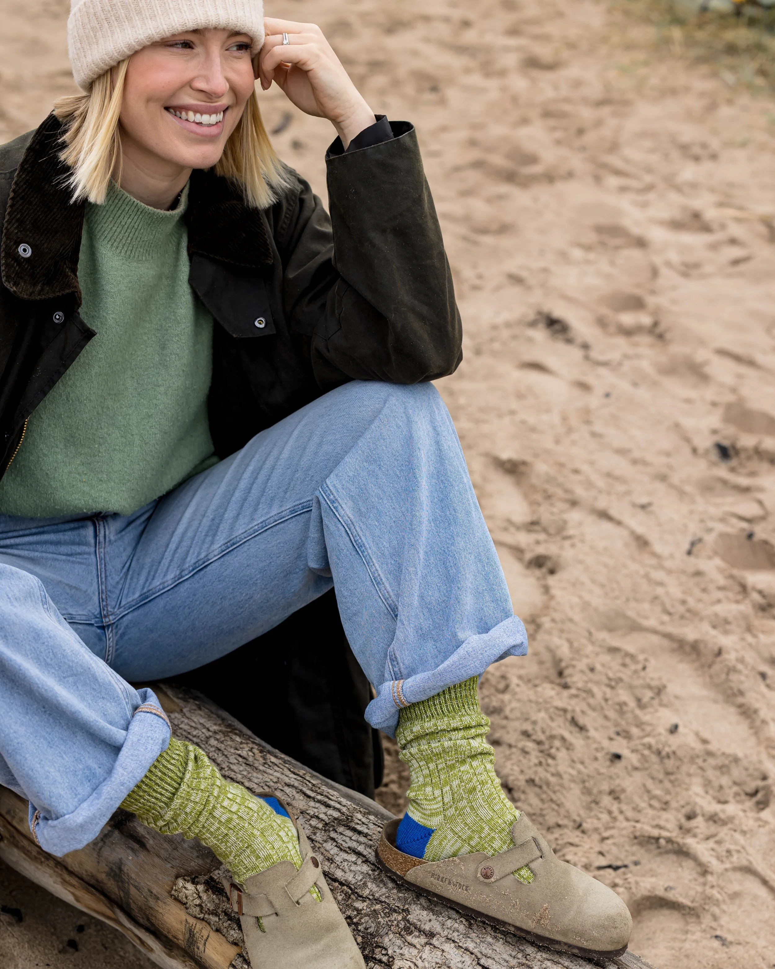 A woman sitting on a log at the beach, smiling, wearing a beige knit hat, green sweater, black jacket, cuffed blue jeans, green patterned socks, and beige shoes.