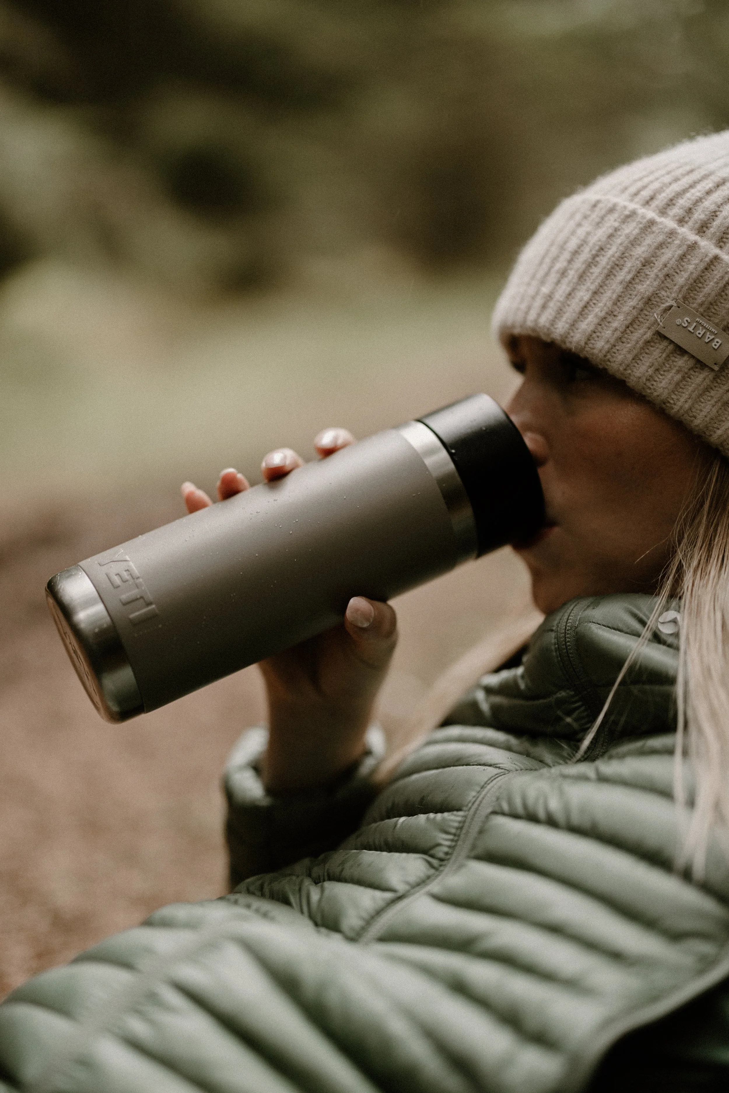 Person wearing a beige beanie and a green puffer jacket drinking from a gray travel mug outdoors.