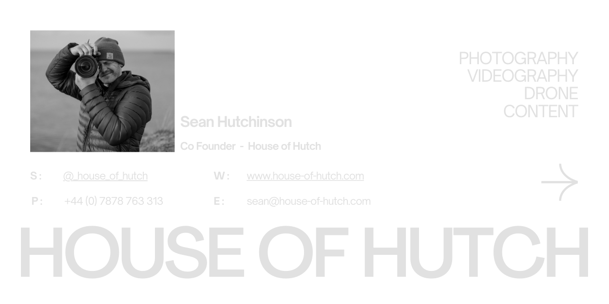 A person in a black jacket and beanie holding a camera, smiling outdoors.